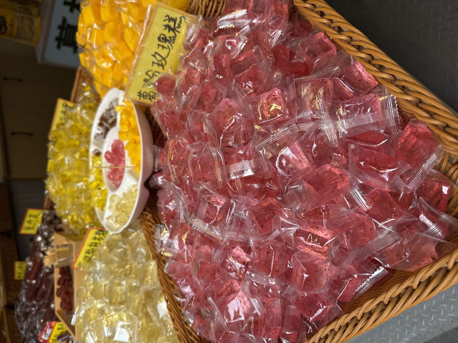 This image depicts a close-up view of a market stall display in Furong, China, featuring various individually packaged candies or jellies. In the foreground, a large wicker basket is filled with numerous small, translucent pink or red square-shaped candies, each sealed in clear plastic packaging. To the left and right background, two additional large wicker baskets hold similarly packaged items; the left basket contains yellow or light-colored candies, and the right basket contains bright yellow candies. A white, multi-compartment dish, positioned centrally in the midground, holds samples of different candies, including pink, yellow, and dark brown varieties, with a few toothpicks visible. Yellow rectangular labels with Chinese characters are placed within the displays, one partially visible on the right, reading "葛粉玫瑰糕" (Géfěn Méiguī Gāo - Arrowroot Powder Rose Cake). The background is blurred, showing hints of other merchandise and shelving, suggesting an indoor retail environment. No people are visible.