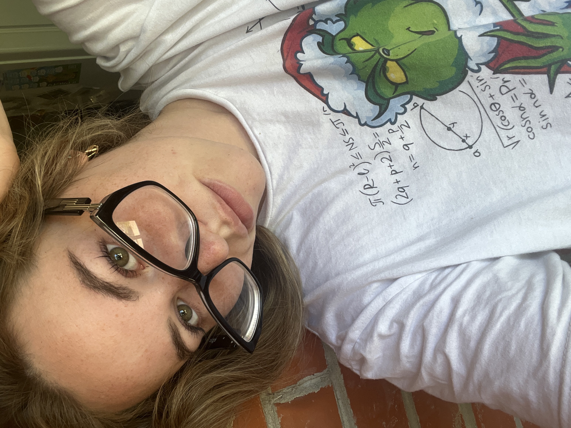 A person wearing black-rimmed glasses and a white t-shirt with a Grinch graphic and mathematical formulas is lying down on a brick surface. The location is not evident.