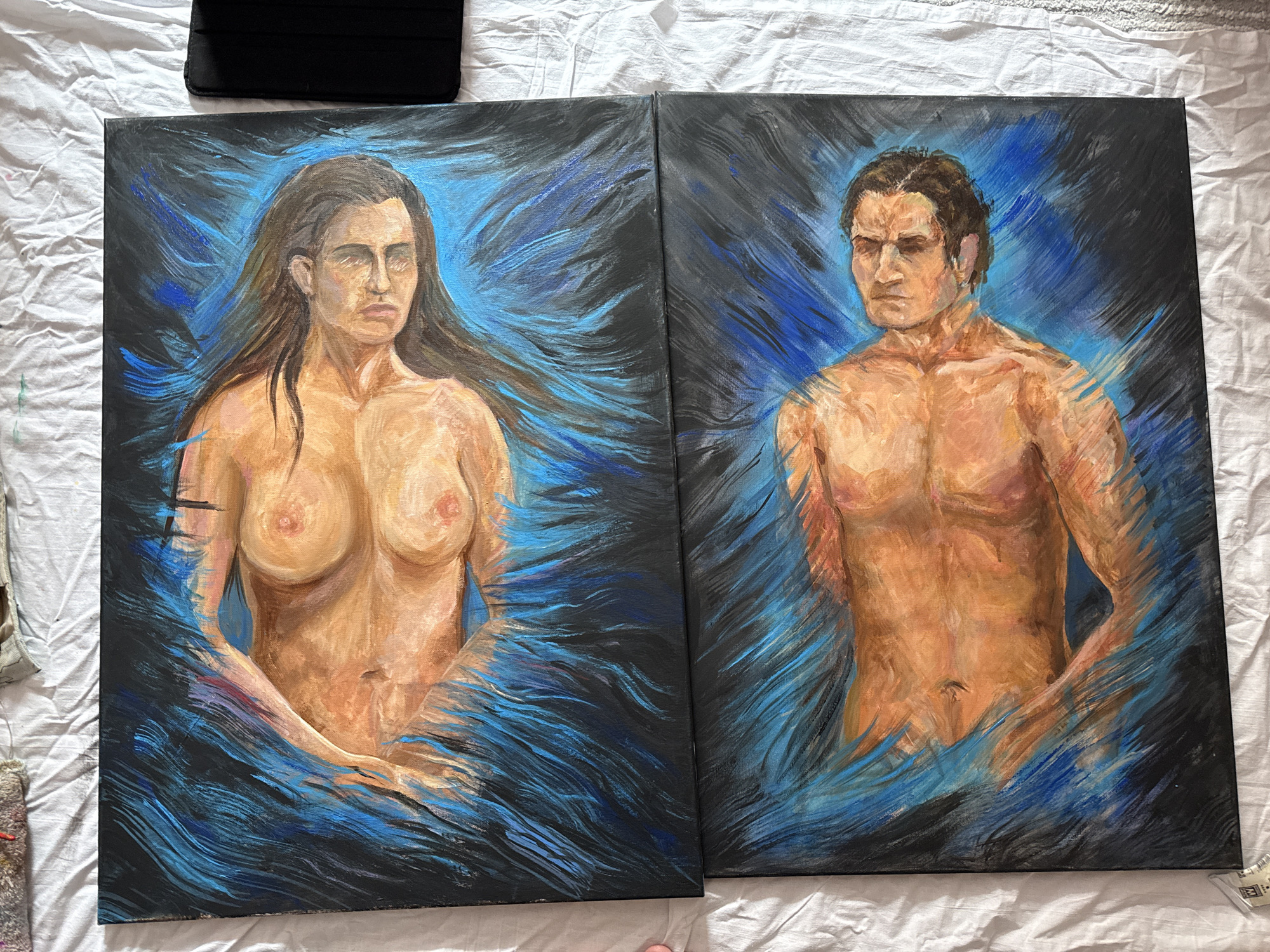 The image shows two oil paintings, side-by-side, depicting a nude woman and a nude man. The woman on the left has long, dark hair and is painted with light brown skin tones. The man on the right has short, dark hair, similarly light brown skin, and muscular features. Both figures appear to be partially submerged or emerging from a swirling mass of black and blue brushstrokes, which constitute the background. The paintings are on separate canvases laid upon a white, crumpled fabric surface. A black tablet case sits atop the painting of the woman and a small tube of paint is in the bottom right corner of the painting of the man. The location is unknown.