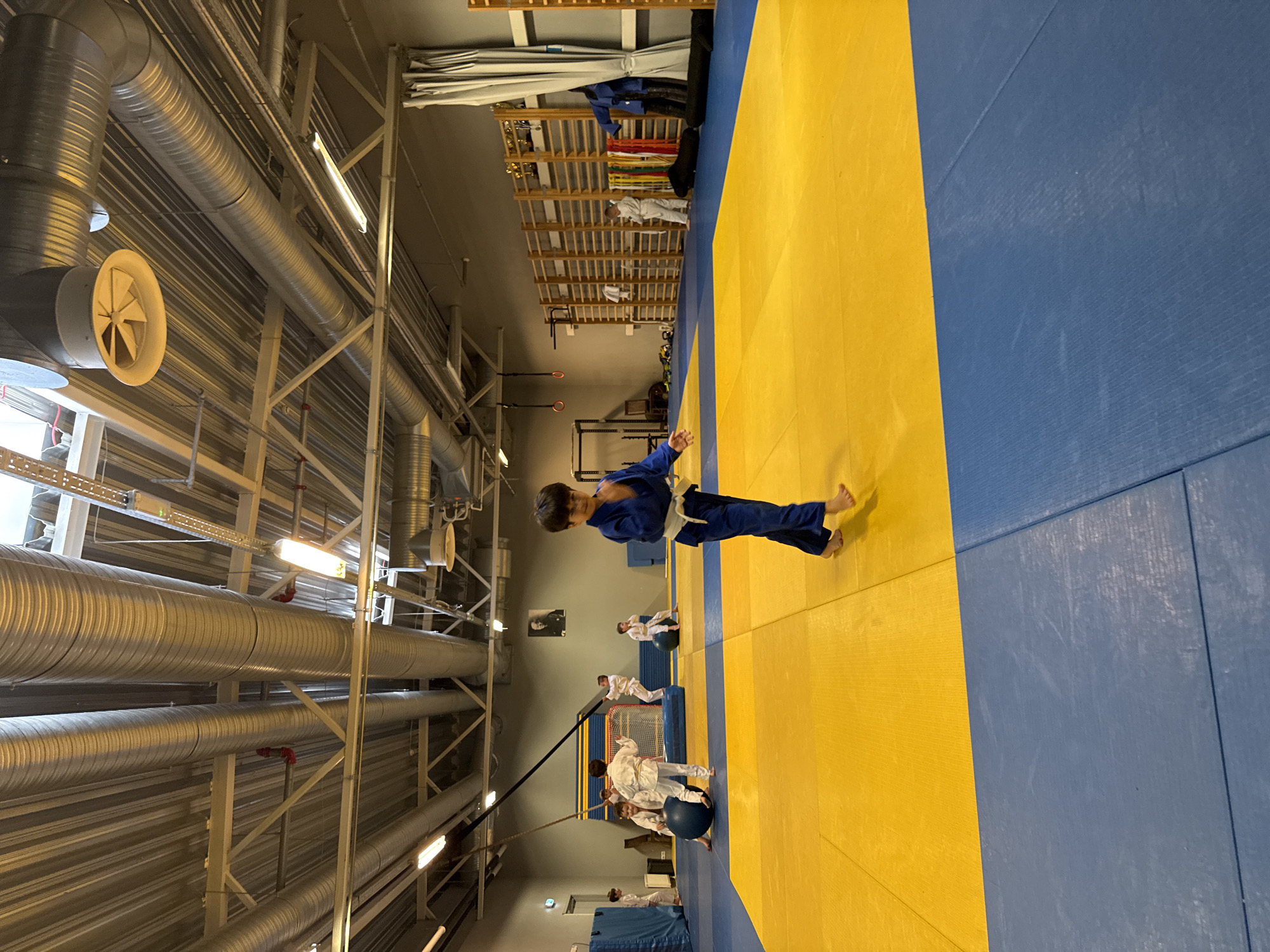 This is a full-shot, eye-level photograph taken indoors of a judo training session. The room has a high ceiling with visible ventilation pipes and fluorescent lighting. The floor is a mat with alternating sections of yellow and blue. In the foreground, a young boy in a blue judo gi walks across the yellow mat, looking towards the camera. In the middle ground, several other children in white judo gis are engaged in activities on the mat, including one sitting on a large blue ball and others standing. In the background, there is a wall with wooden shelves displaying various items, a large grey curtain, and a framed picture of a man on the wall. The overall environment appears to be a sports hall or gymnasium dedicated to martial arts training. The image location is the Tallinn, Estonia sports hall.