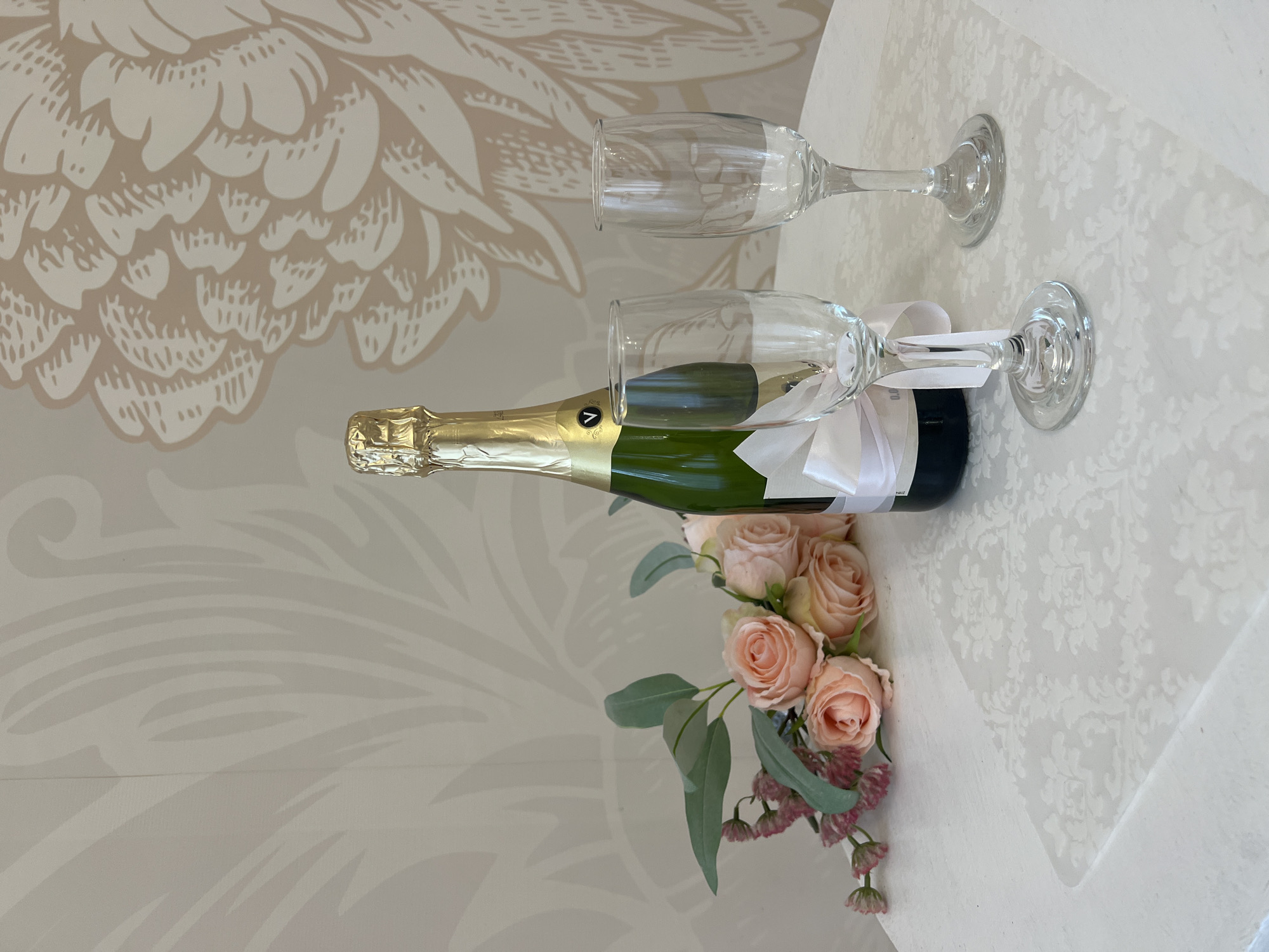 An overhead shot captures a celebratory setup on a light-colored table in Kyiv, Ukraine. The scene features a green glass bottle of sparkling wine or champagne with a gold foil top and a gold label, partially obscured by a clear champagne flute. This flute has a white ribbon tied around its stem, which also appears to secure it to the bottle. A second, unadorned clear champagne flute stands beside it.
To the left of the bottle and glasses lies a small, elegant bouquet of flowers. It consists primarily of several light pink or peach roses, accented with long, silvery-green eucalyptus leaves and clusters of tiny, reddish-pink flowers.
The items rest on a white or light cream-colored tablecloth that features a subtle, embossed or woven damask-like pattern across its surface. Behind the table, a wall is visible, decorated with a large, stylized floral or feathered pattern in a slightly darker, muted beige tone. The lighting is soft and even, suggesting an indoor setting. The arrangement implies an intimate celebration or special occasion for two. On the champagne bottle's label, a distinct white 'V' logo is visible, along with some unreadable black text.