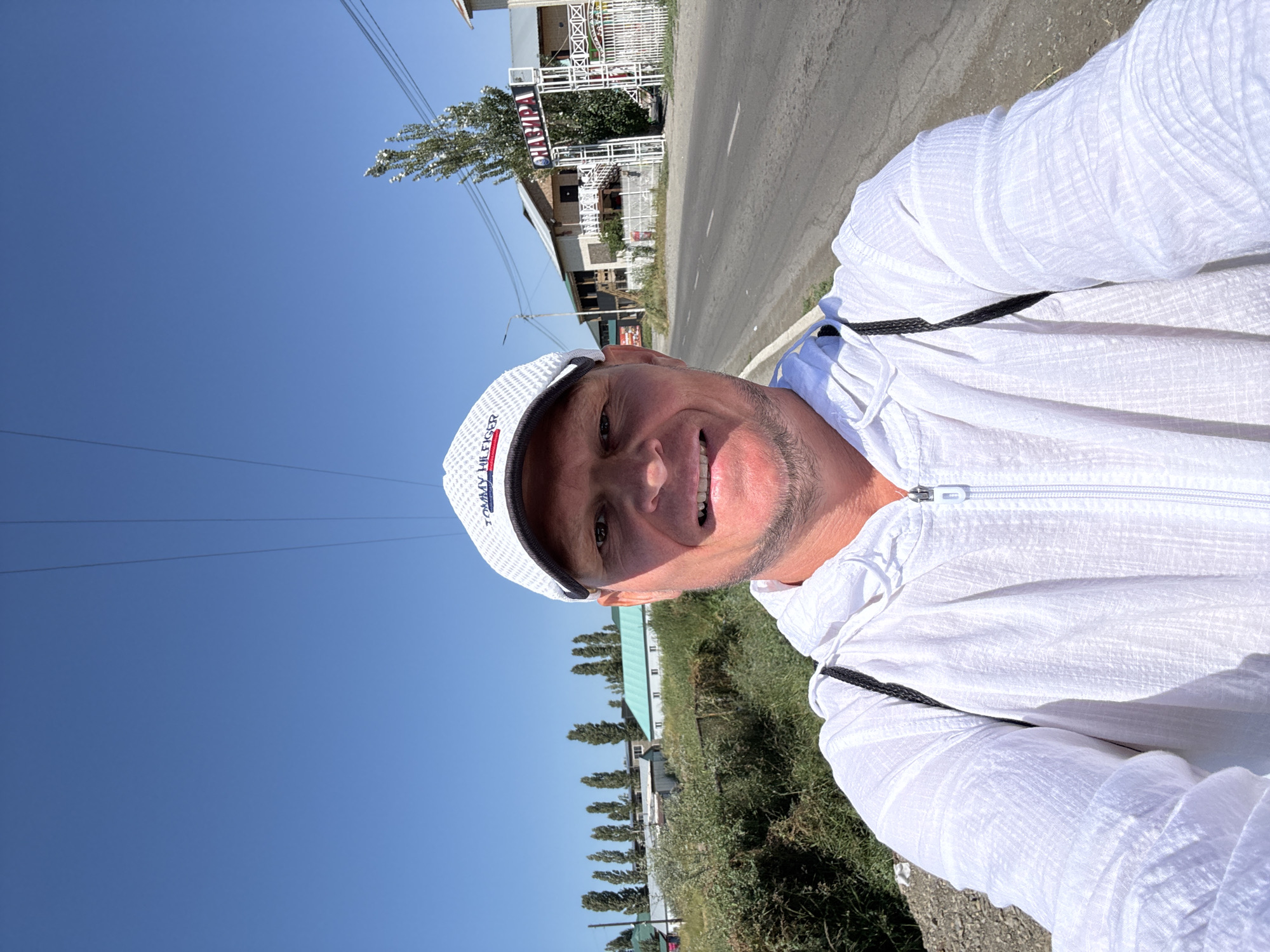 A man in a white hooded jacket and a white baseball cap takes a selfie outdoors. The man is fair-skinned with short-cropped hair and a light beard. He is smiling with his teeth showing. The background is a street scene in the city of Zharbulak, Kazakhstan. On the right side of the image, there is an archway with the word "CHAYKHANA" written in Cyrillic letters, indicating a tea house or restaurant. Buildings and trees line the street. The sky is clear and blue.