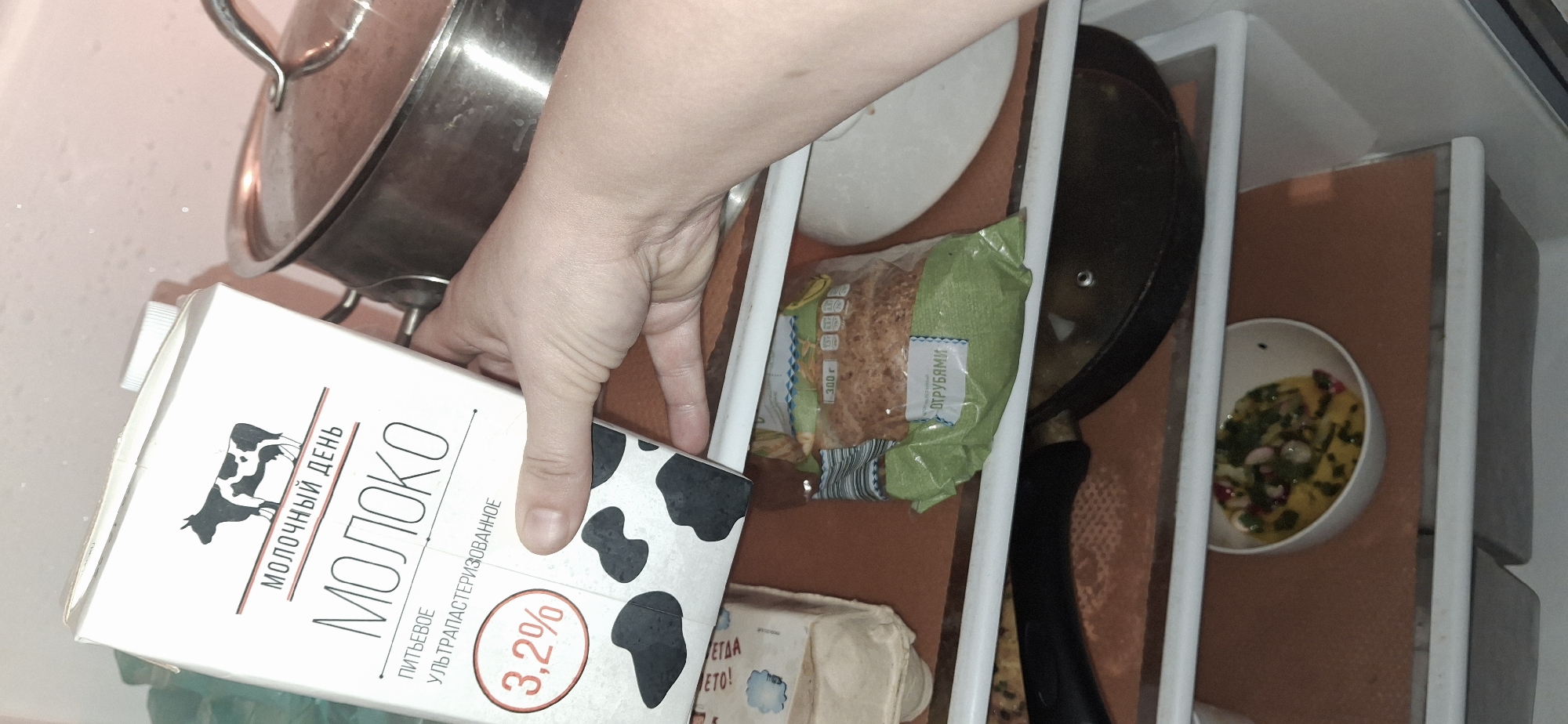 A human hand is holding a carton of milk in a refrigerator, partially obscuring a stainless-steel pot. The milk carton is white with black cow spots and text in Russian including "МОЛОКО" and "3.2%". The refrigerator interior contains other items including a covered frying pan, a bowl of food, a package of something, and eggs. The image appears to have been taken inside a refrigerator in Писарёвка, Russia.
