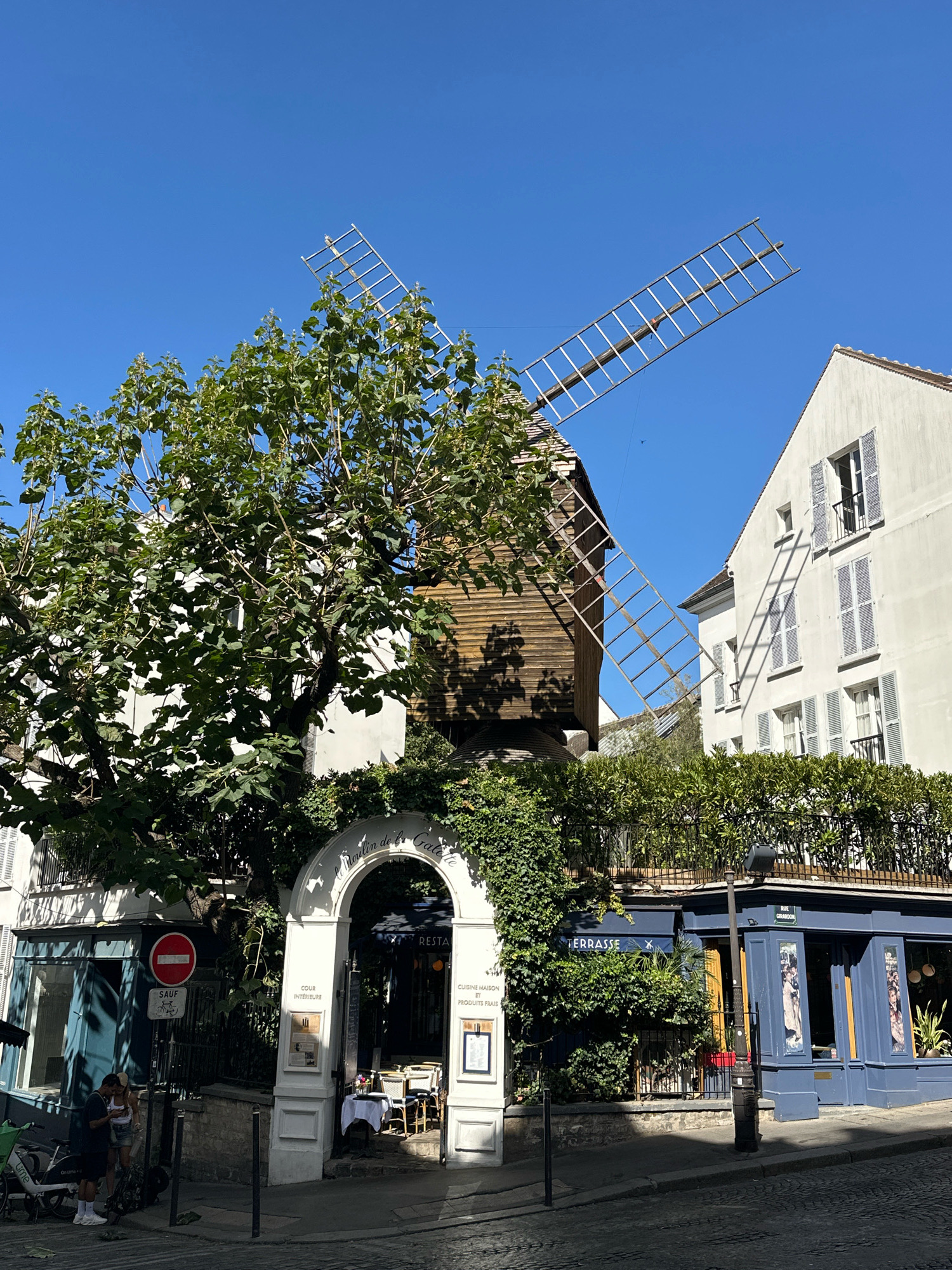 The image captures a street scene in Montmartre, Paris, France, on a clear, sunny day. Dominating the left side of the frame is a weathered wooden windmill, its large, bare wooden sails extended diagonally against the bright blue sky. Lush green foliage from a large tree partially obscures the windmill's base and one of its sails.
To the right of the windmill, a white building with traditional grey shutters on its windows is visible. A shadow of the windmill's sails is cast across its facade, creating a striking geometric pattern.
In the foreground, a white arched entryway leads to an interior courtyard. The archway is adorned with ivy and the sign "Le Moulin de la Galette" is visible. Inside the courtyard, several tables with white tablecloths are set for dining.
Below the archway, a blue facade building with "TERRASSE" signage is visible, suggesting it is a restaurant or cafe with outdoor seating. A black lamppost stands in front of this building.
On the left side of the entryway, a blue building with large windows is seen, with a "SAUF" (except) sign and a red circular "no entry" sign displayed on a post. Two individuals, a man and a woman, are standing near the street on the left, facing a bicycle. The street itself is paved with cobblestones, indicating a historic Parisian district. The overall atmosphere is that of a charming, sun-drenched street in a well-known Parisian neighborhood.