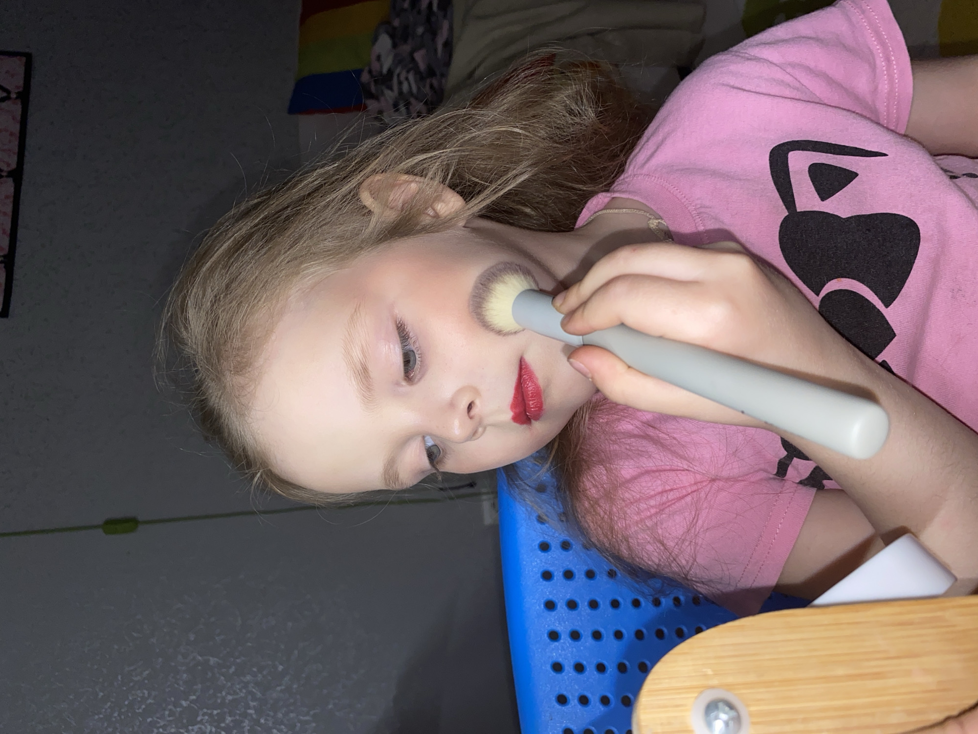 A young child, likely a girl with light hair, fair skin, and blue eyes, is depicted engaged in playing with makeup in an indoor setting. She is seen applying blush or powder to her left cheek with a makeup brush, which she holds with her right hand. Her lips are already adorned with somewhat messily applied bright red lipstick.
She is wearing a short-sleeved pink t-shirt featuring a black graphic design, possibly a heart or an abstract shape. The child appears to be reclining or leaning back against a blue plastic chair or seat, which has a textured, perforated surface. Part of a light-colored wooden furniture piece, possibly an armrest or table, is visible in the foreground.
The background consists of a dark wall. Hints of other elements are visible: a section of a rainbow-colored item, possibly a flag or decoration, is seen in the upper right background, partially obscured by a dark, patterned fabric. A narrow green cord or wire runs along the wall on the left. The overall lighting is dim, suggesting an indoor environment, possibly in the evening. The child's expression conveys focus and engagement in her playful activity. No discernible text is visible in the image. The location context provided is Votkinsk, Russia.