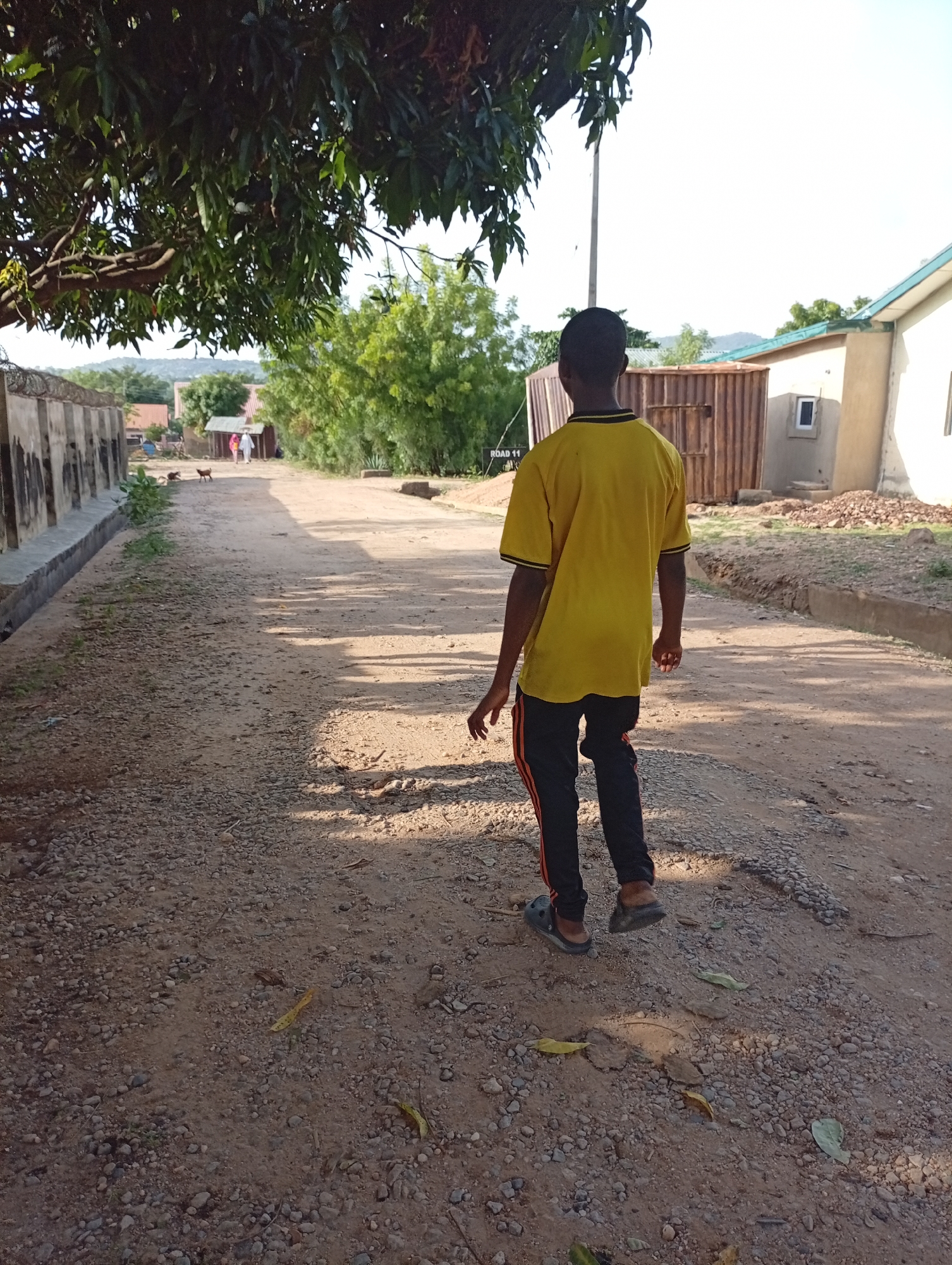 A photograph taken in Adankolo, Nigeria depicts a person walking away from the camera on a dirt road. The person is wearing a yellow shirt, dark pants with orange stripes, and sandals. The road appears unpaved and uneven. To the left of the road is a fence, and to the right, a partially visible building. Trees overhang part of the road, providing shade. Further down the road, there are glimpses of other buildings and greenery.