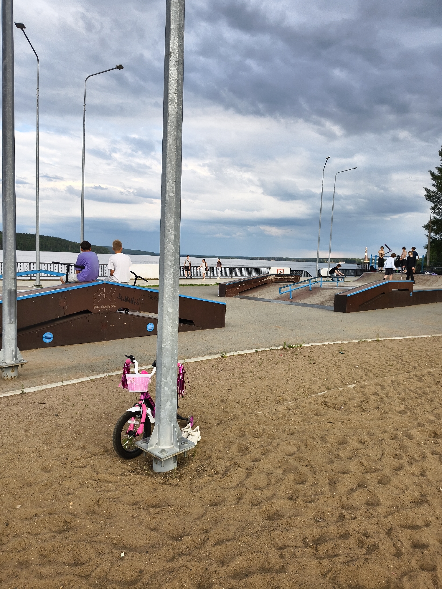 This image depicts an outdoor skatepark located on the shore of the Kama River in Chaikovsky, Russia. The foreground features a sandy area with a pink children's bicycle adorned with streamers and a basket parked near a metal pole. In the mid-ground, a complex of wooden ramps and obstacles forming the skatepark is visible. Several young people are present, some observing from benches on a raised embankment, while others are actively engaged in skateboarding and scootering on the ramps. The background reveals the expansive Kama River, a tree-lined shore, and a cloudy sky overhead. The scene captures a moment of leisure and recreation in a waterside urban environment.