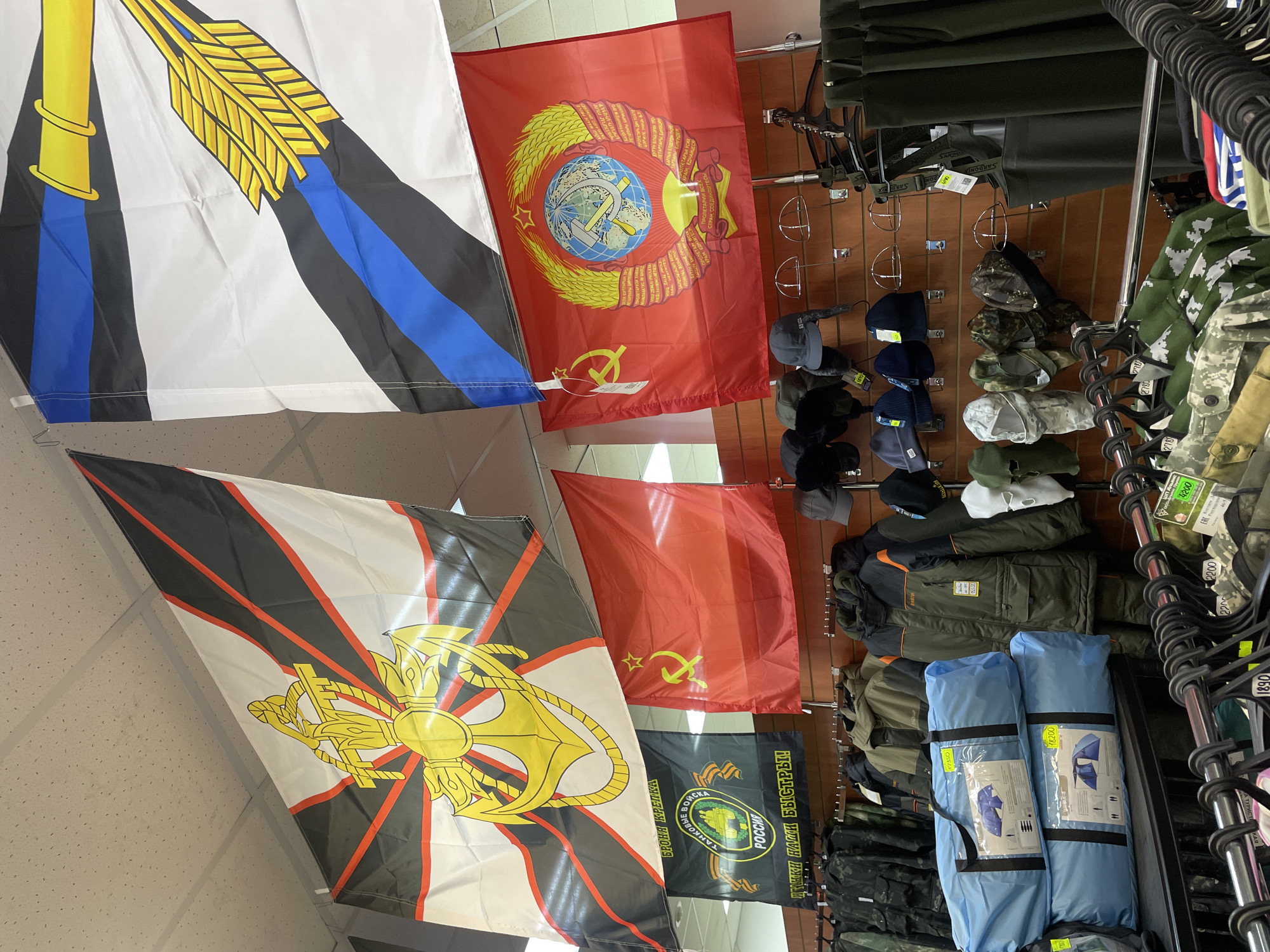 This image captures a retail display in Krasnoyarsk, Russia, featuring a variety of military-themed and outdoor merchandise alongside several flags.
Hanging from the ceiling are multiple large flags. To the top left, a white flag with horizontal black and blue stripes features a prominent golden emblem resembling a staff or column topped with a sheaf of wheat or feathers. Next to it is a red flag bearing the full emblem of the Soviet Union, including the hammer and sickle, a globe, and a wreath of wheat, with the motto "Proletarians of all countries, unite!" written in various languages around the wreath. Below these, a white flag with radiating black and red stripes carries a large golden emblem of a fouled anchor crossed with two axes or sabers, identifying it as a flag associated with the Russian Navy. Another red flag with a simpler golden hammer, sickle, and star is visible further down. A black flag for the Russian Tank Troops is also displayed, featuring a green circular emblem with a tank and a red star, along with Cyrillic text including "БРОНЯ КРЕПКА" (Armor is strong), "ТАНКОВЫЕ ВОЙСКА" (Tank Troops), "РОССИЯ" (Russia), and "ТАНКИ НАШИ БЫСТРЫ!" (Our tanks are swift!).
On the right side of the image, various items are displayed on wall-mounted pegboards and racks. These include military-style hats and caps in camouflage patterns and dark colors, as well as several jackets and outerwear. Two blue camping tents, still packaged in their bags, are stacked on a lower shelf. Price tags are visible on many items, with numbers like "17550", "18200", "2200", "1890", "5500", "4200", "278", and "2780," likely indicating prices in Russian Rubles. Some product labels include "SARDONI" and "SARD MIX." The setting appears to be an indoor store, with white ceiling tiles and wooden paneling on the walls.
