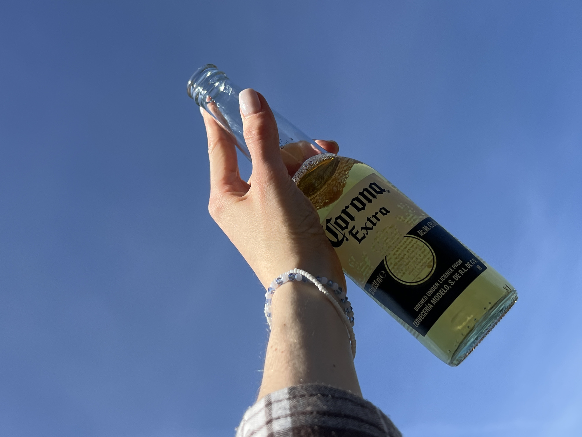 The image shows a person's arm holding a Corona Extra beer bottle against a clear blue sky. The person is wearing two bracelets on their wrist, one white and beaded and the other with blue and white beads. The arm is clad in a plaid shirt. The bottle is held at an angle, showcasing the label with "Corona Extra" prominently displayed. The bottle contains a pale yellow liquid, likely beer. The background consists solely of the bright blue sky. The location is not specified.