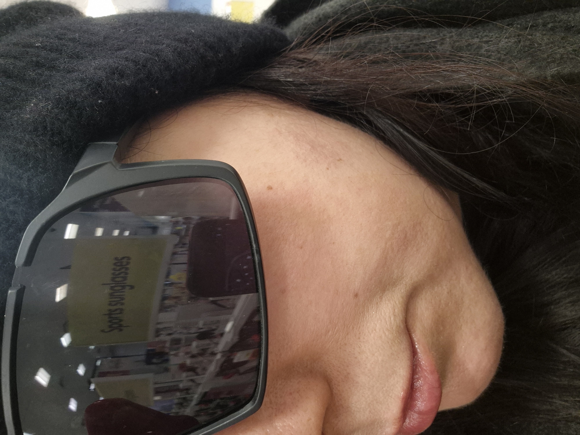 A close-up, rotated image shows a person wearing sunglasses. The reflection in the sunglasses shows a store display with a sign reading "Sports Sunglasses." Given that the question specified the city of West Suffolk, United Kingdom, a reasonable assumption is that the photograph was taken in a retail establishment within this city that sells sunglasses. The person has dark hair and skin. The photograph focuses primarily on their face, specifically the eye and nose area obscured partially by the sunglasses. A black textured material (possibly a hat or clothing) is seen near the top of the head. The lips are visible and appear slightly glossy.