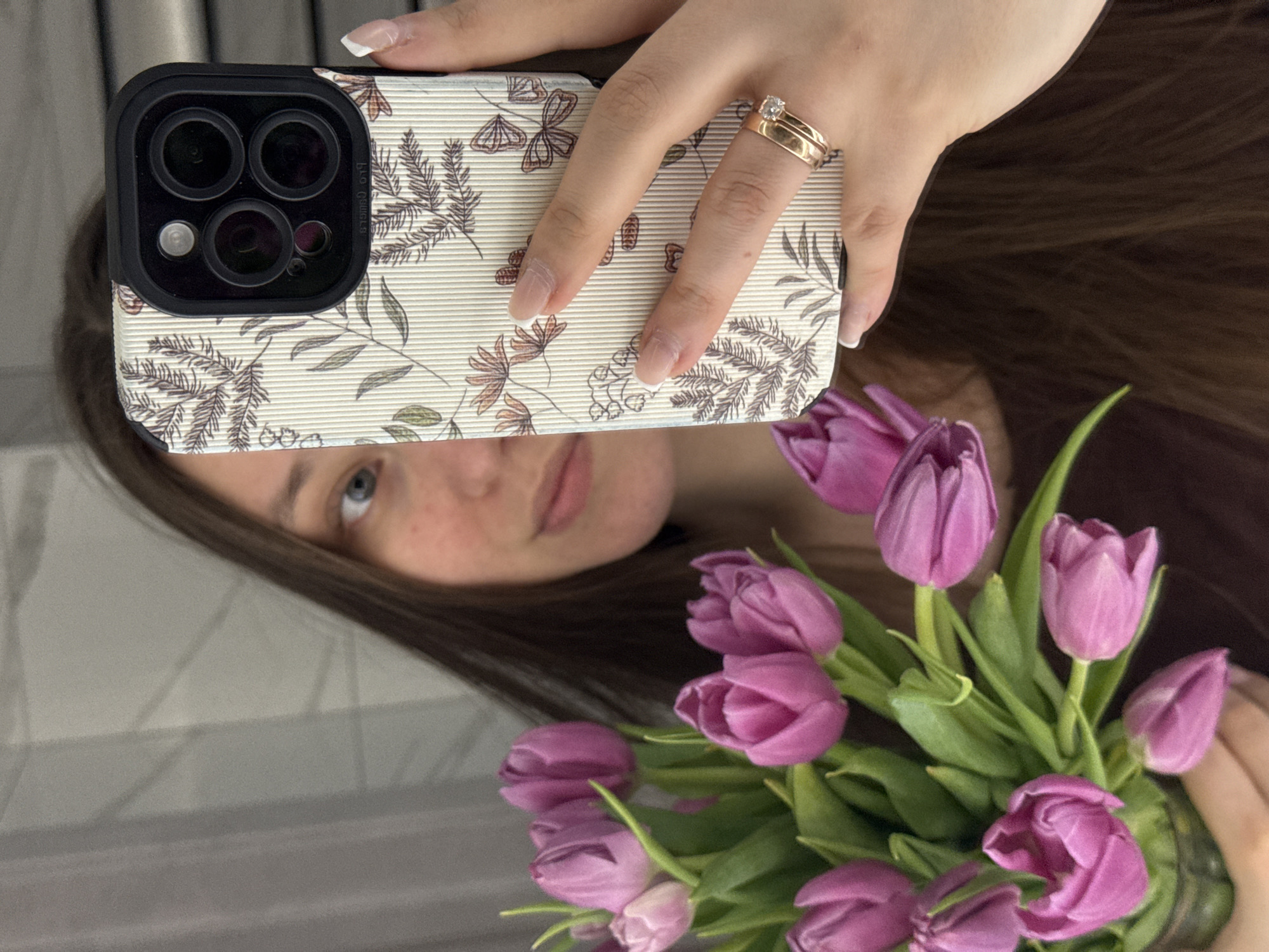 The image displays an indoor mirror selfie taken in Krasnodar, Russia. A female subject, with long dark brown hair and blue eyes, is partially visible in the reflection, with her face largely obscured by a smartphone. She is holding an iPhone, identifiable by its three-lens camera system, encased in an off-white, vertically ribbed protective case decorated with a botanical pattern of brown leaves, orange flowers, and butterflies. Her right hand, featuring French-manicured nails, grips the phone, and two gold rings—one plain band and one with a single clear stone—are worn on her ring finger. In her left arm, she holds a bouquet of approximately 10-12 vibrant purple tulips with green stems and leaves. The background, visible in the reflection, suggests a light-colored, subtly textured wall or surface consistent with a domestic interior.