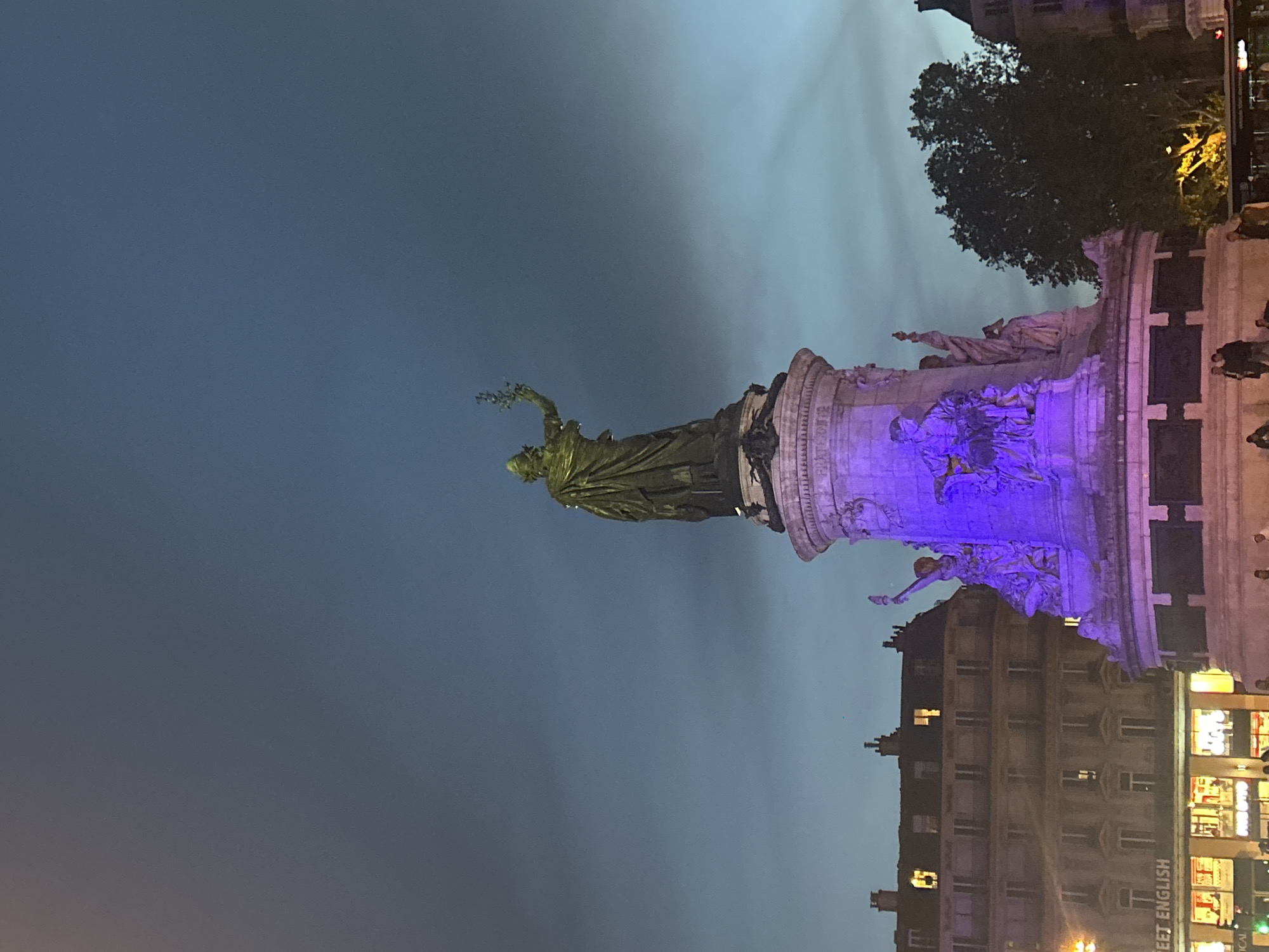 The image is a nighttime shot of the Place de la République in Paris, France. The central feature is the statue of Marianne, a symbol of France, standing atop a large pedestal. The statue and the pedestal are illuminated with purple light. A dark, cloudy sky forms the backdrop. Buildings are visible around the square, including what appears to be a commercial building. There are people milling around the base of the monument. A tree is to the right of the monument.