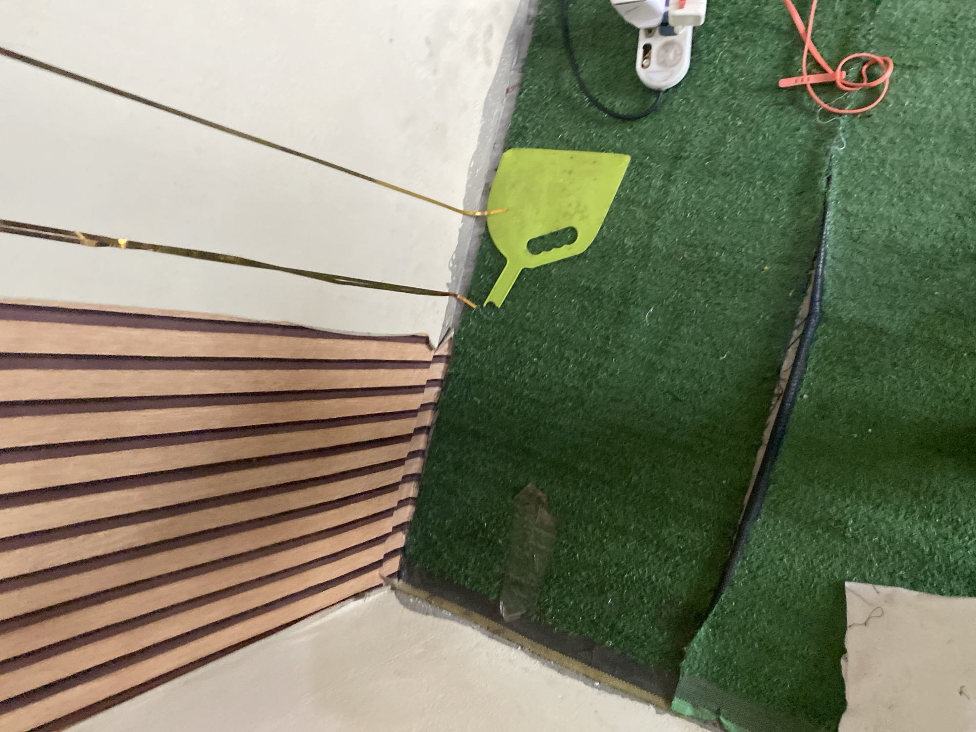 The image depicts a close-up, high-angle view of an indoor scene with artificial turf covering the floor. To the left, a section of wall is paneled with vertical wooden slats, alternating between a light tan and a dark brown color. The artificial turf is green and appears to be laid out in two sections, with a visible seam or edge between them.
On the turf, several objects are present. A clear plastic bottle, possibly crushed, lies on the turf in the lower left portion of the image. A bright lime green, flat, shovel-like object with a handle is suspended by two thin, dark-colored strings. The strings appear to be attached to a higher point, out of frame. Further to the right, a white object with red and black accents, resembling an electronic device or a tool, is visible. A coiled bright orange cord or rope is also on the turf to the right of the white device.
The lighting in the image is somewhat diffused, suggesting an indoor environment with artificial light or indirect natural light. The background wall is a light, neutral color. There are no people visible in the image. The scene appears to be domestic or a recreational space, possibly a home or a small training area. The specific location in Jauro Yinu, Nigeria, cannot be determined from the visual information provided in the image alone.