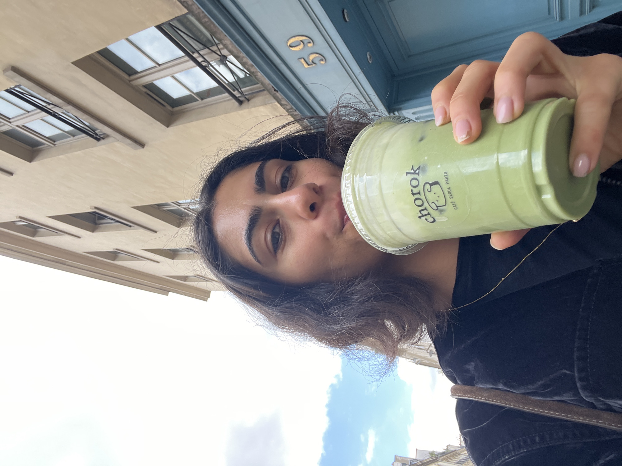 A woman is taking a selfie while drinking a green beverage from a clear plastic cup with a white lid. The cup has the text "chorok Cafe Seoul Paris" and a cartoon bear on it. She is holding the cup with her right hand. She has dark hair and is wearing a dark-colored jacket. Behind her is a building with windows and a blue door with the number 69. The location is Paris, France.