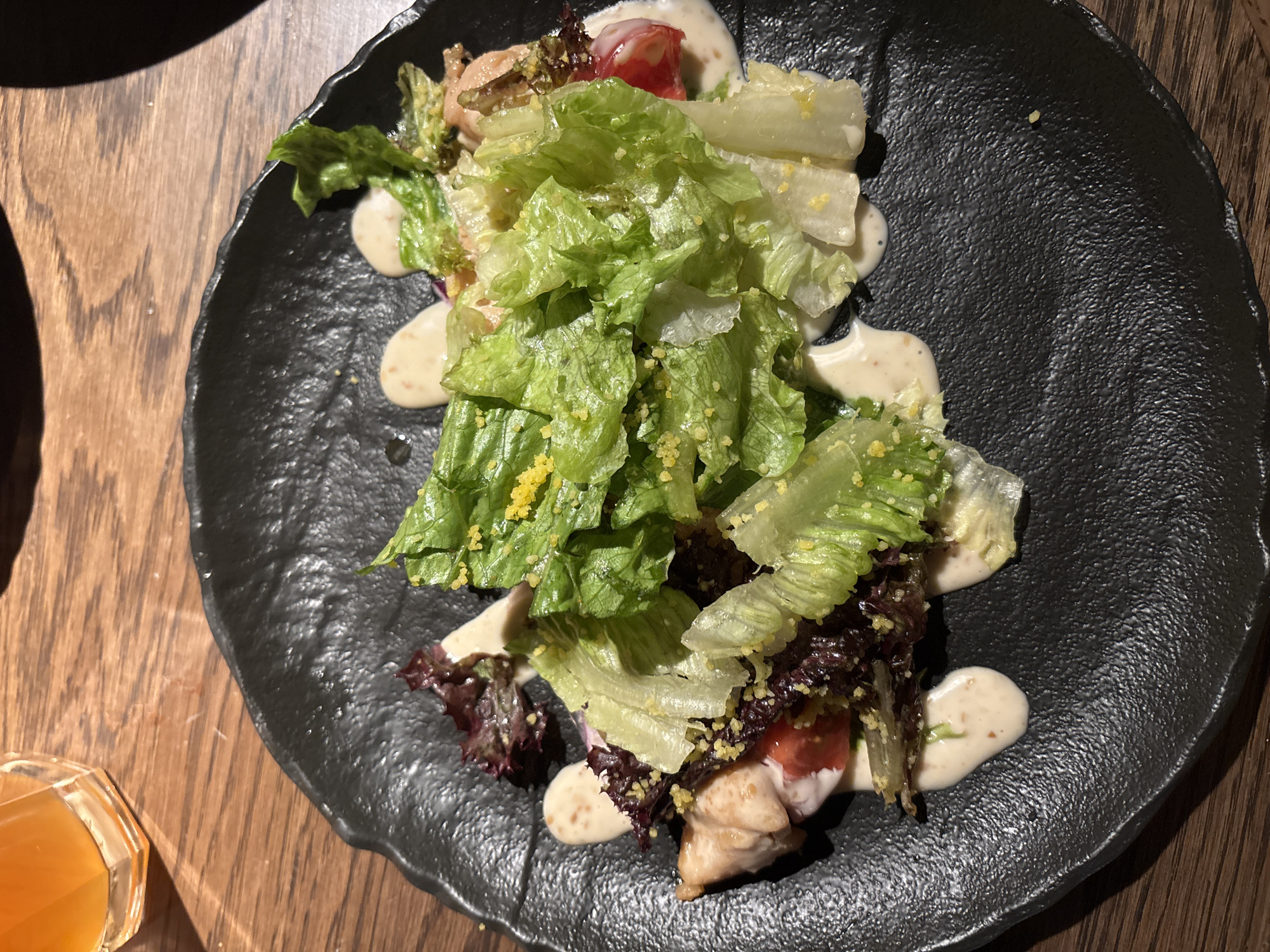 A top-down shot depicts a salad on a textured black plate, positioned on a wooden surface. The salad includes a mix of green and red lettuce leaves, sliced tomatoes, and what appears to be chicken or fish pieces. A creamy dressing is drizzled throughout the salad. A glass of orange liquid is partly visible in the lower-left corner of the frame. There are no people depicted in the image, and the environment suggests an indoor setting, possibly a restaurant. There is no information available to establish the location as Moscow, Russia.