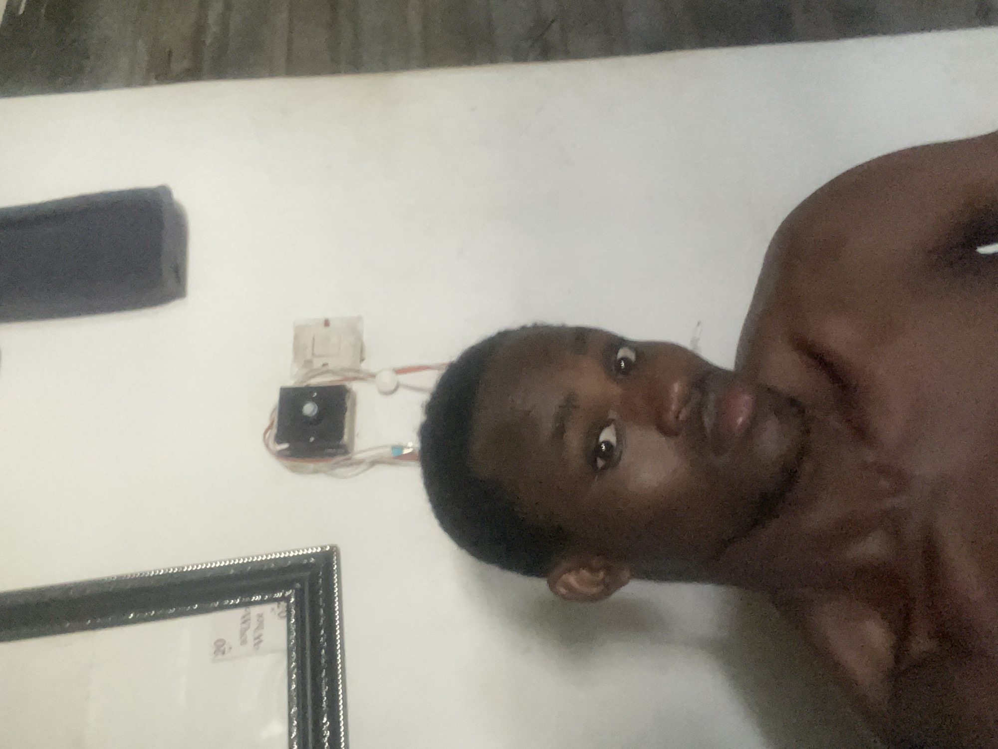 The media file is a close-up, vertically oriented photograph of a person. The subject is a young Black male with dark, short hair and brown eyes. He appears to be looking directly at the camera with a neutral expression. His bare chest is visible in the foreground.
The background is a plain white wall. On the wall, to the left of the subject's head, there is a black rectangular object mounted horizontally. Below this object, there is a white electrical outlet or junction box with wires connected to it. Further to the left, in the lower corner, a framed picture or certificate is partially visible, with a patterned silver frame. Some faint text is visible on a white label within the frame, including the number "20". The lighting in the image is somewhat dim, suggesting an indoor setting, possibly at night or in a room with limited natural light. There are no clear indicators of time of day or weather. The overall impression is a candid or personal snapshot.