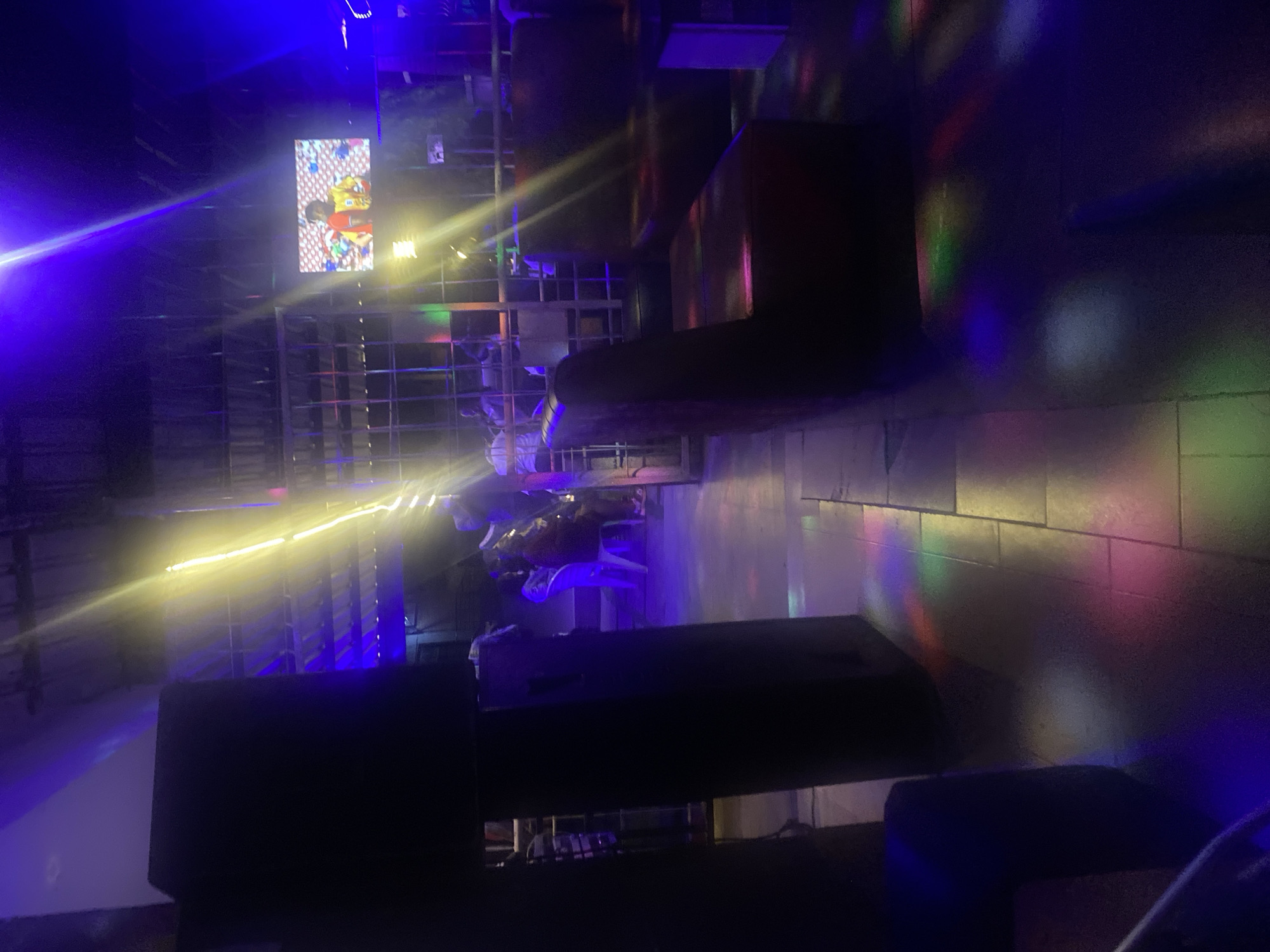 This image depicts an interior view of an entertainment venue, likely a bar or lounge, situated in Jos, Nigeria. The scene is dimly lit by artificial sources, primarily dominated by blue and purple ambient lighting originating from overhead structures, which appear to include exposed beams or a corrugated ceiling. Several strong, elongated streaks of yellow light traverse the upper and central portions of the frame, suggesting decorative string lights or illuminated structural elements.
On the left side of the foreground and midground, two tall, dark, rectangular objects, likely large audio speakers, stand vertically. Padded, dark reddish-brown booth seating is arranged along the right side of the venue, with some sections appearing unoccupied. In the midground, towards the left, white plastic chairs are visible, occupied by indistinct figures. Several individuals are present throughout the midground and background, mostly seated or standing, their specific features obscured by the low light conditions. One person is partially visible on the far left in a white top.
A brightly illuminated television screen is mounted on a far wall in the upper-right quadrant, displaying a still image of an individual wearing a red and yellow top against a patterned background. Metal grating or railing structures are visible vertically in the midground, serving as partitions or structural supports. The tiled floor reflects a kaleidoscope of red, green, blue, and yellow light patterns, indicative of a disco lighting effect. No explicit actions or clear interactions between individuals are discernible.