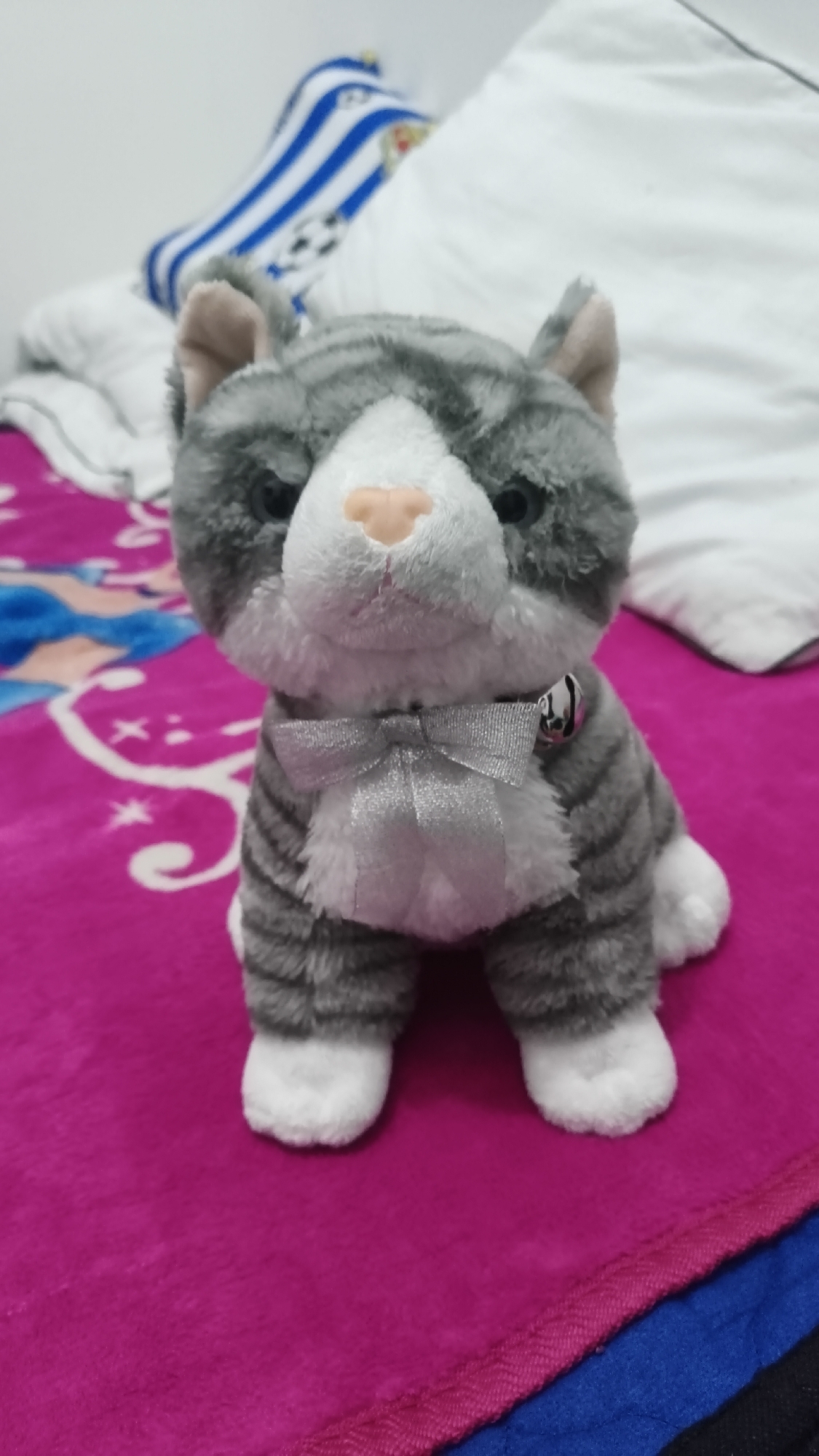 A stuffed cat toy with gray and white fur, a pink nose, and a silver bow is sitting on a fuchsia blanket. The cat toy also has a small silver bell around its neck. In the background, there are pillows and blankets, one of which has blue and white stripes with a soccer ball pattern. The scene appears to be indoors, possibly on a bed. There is no indication of the location being in Betung, Indonesia, or any other specific city.