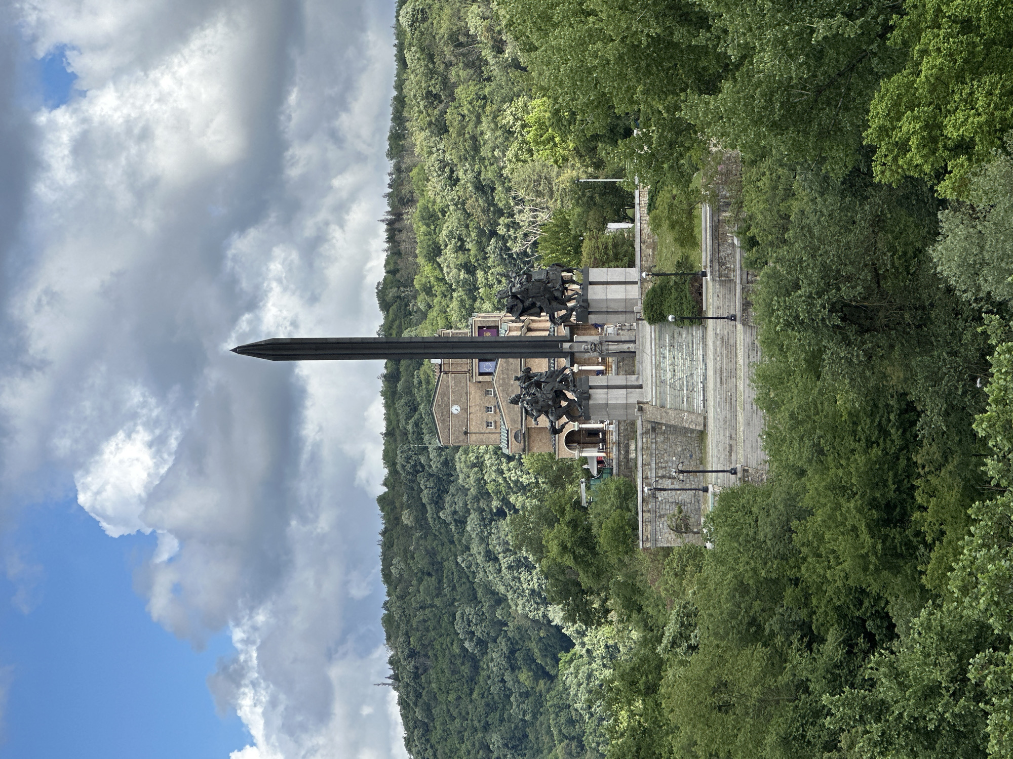 Captured from an elevated perspective, the image features the Asenevtsi Monument in Veliko Tarnovo, Bulgaria, set against a backdrop of a lush, forested hillside and a partly cloudy sky. The monument, composed of a tall, dark obelisk-like structure and several bronze statues of horse-riding figures, is situated on a terraced stone platform. Adjacent to the monument is a building with a clock face visible. The stairs leading to the monument are also prominently featured. The foreground and background are dominated by dense green trees, creating a frame around the monument. No people are distinctly visible within the scene.