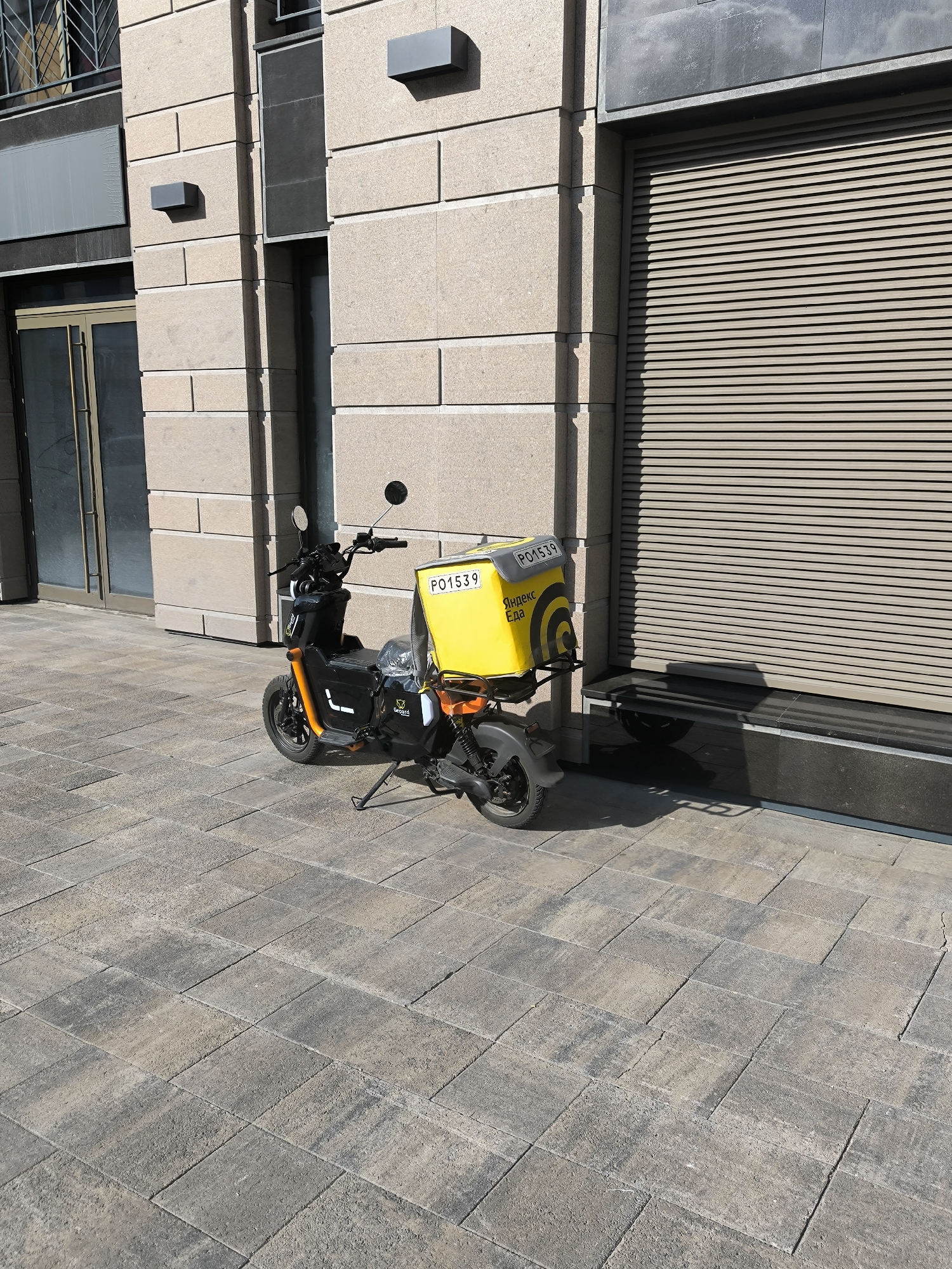 A black and orange electric scooter with a yellow delivery box is parked on a stone-paved sidewalk in front of a building with a beige facade and a large, beige, louvered door or window covering. The scooter has a license plate that reads "PO1539" and branding that includes the Yandex.Eda logo. The building appears to be modern, with some dark gray architectural elements and what look like sconces on the wall. The lighting suggests it is daytime, with shadows cast on the pavement. The environment is urban, with the scooter likely waiting for or having completed a delivery. The background of the building shows some architectural details like windows and a metal grate.