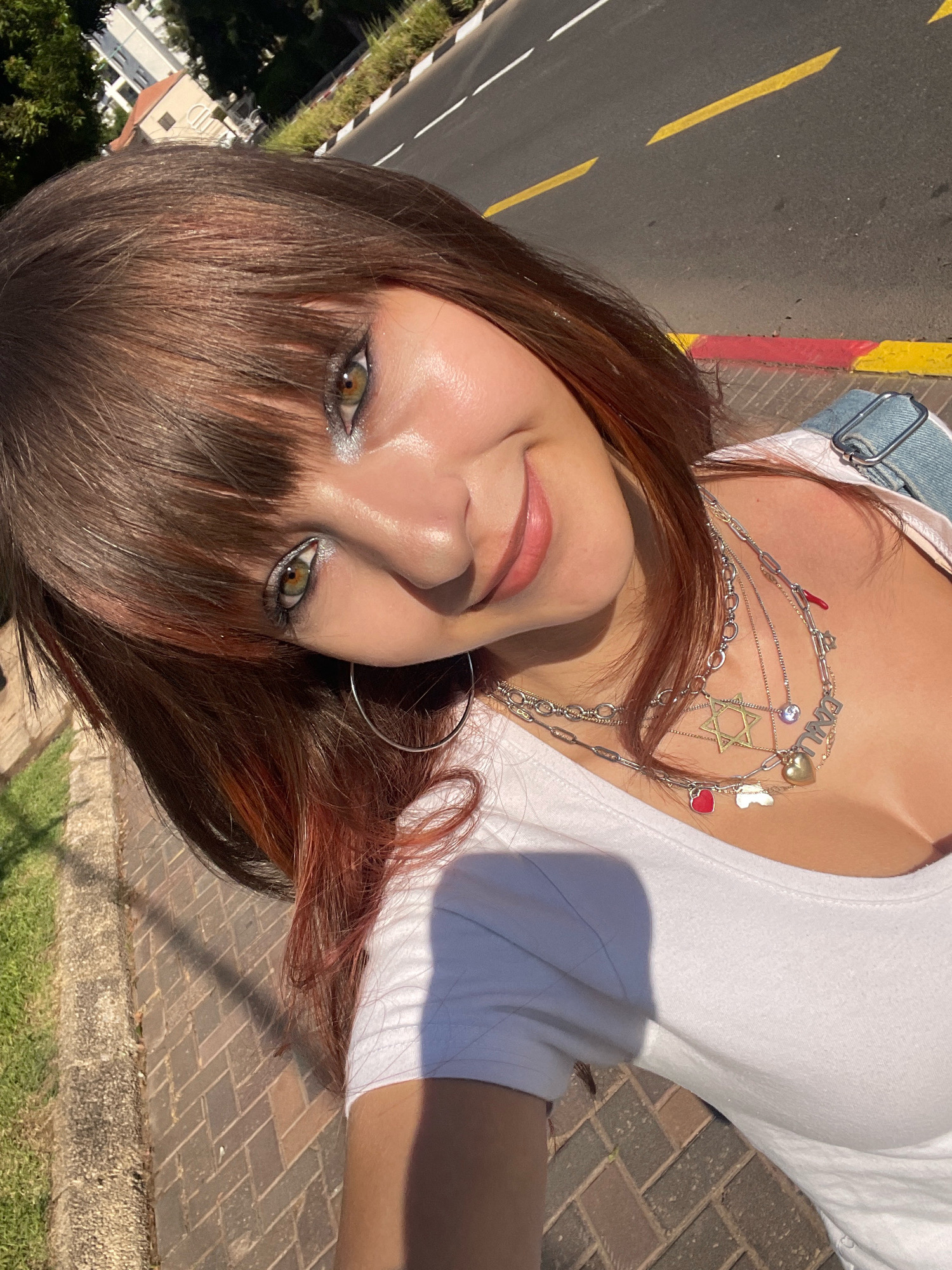 A young woman with brown hair, streaked with reddish tones, is taking a selfie outdoors in Raanana, Israel. She is wearing a white t-shirt and denim overalls. Her neck is adorned with several layered necklaces, including one with a Star of David pendant, a heart charm, and a charm that appears to be a cloud. She has makeup on her eyes, including glittery eyeshadow. The background shows a paved sidewalk, a grassy area, a road with yellow and red markings, and buildings in the distance. The lighting suggests it is a sunny day.