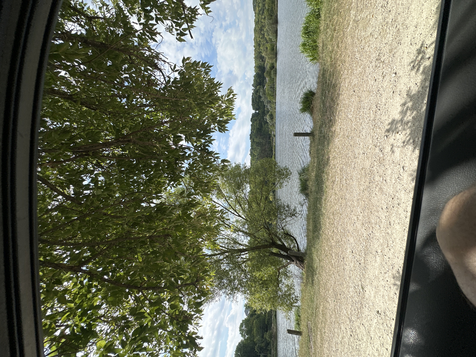 The image shows a view from inside a vehicle looking out at a body of water, likely a lake or river, with trees and a wooded shoreline in the background. The foreground is a gravelly bank, and some of the vehicle's interior, including a person's leg, is visible at the bottom of the frame. The sky is partly cloudy. The location is identified as being in Makiv, Ukraine.