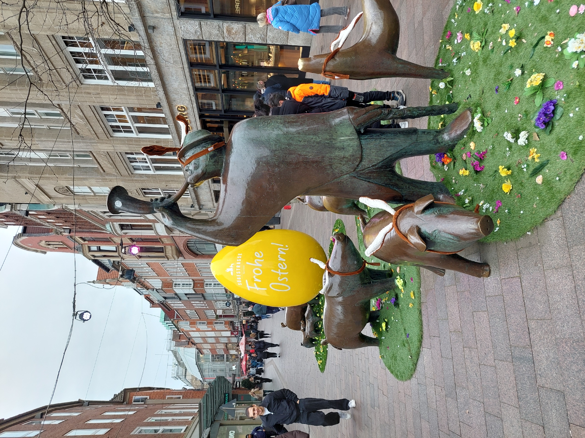The image depicts a scene on a pedestrianized street in Stadtgebiet Bremen, Germany. A bronze statue of a man playing a horn, with bronze animals pulling a carriage and a large yellow Easter egg beside it is visible on a section of green artificial turf adorned with flowers. The egg has the words "Frohe Ostern" written on it. People are seen walking along the street in the background. Buildings line both sides of the street, and street lights are hanging overhead. A man is standing near the front of the statue looking at the camera. The weather appears overcast.