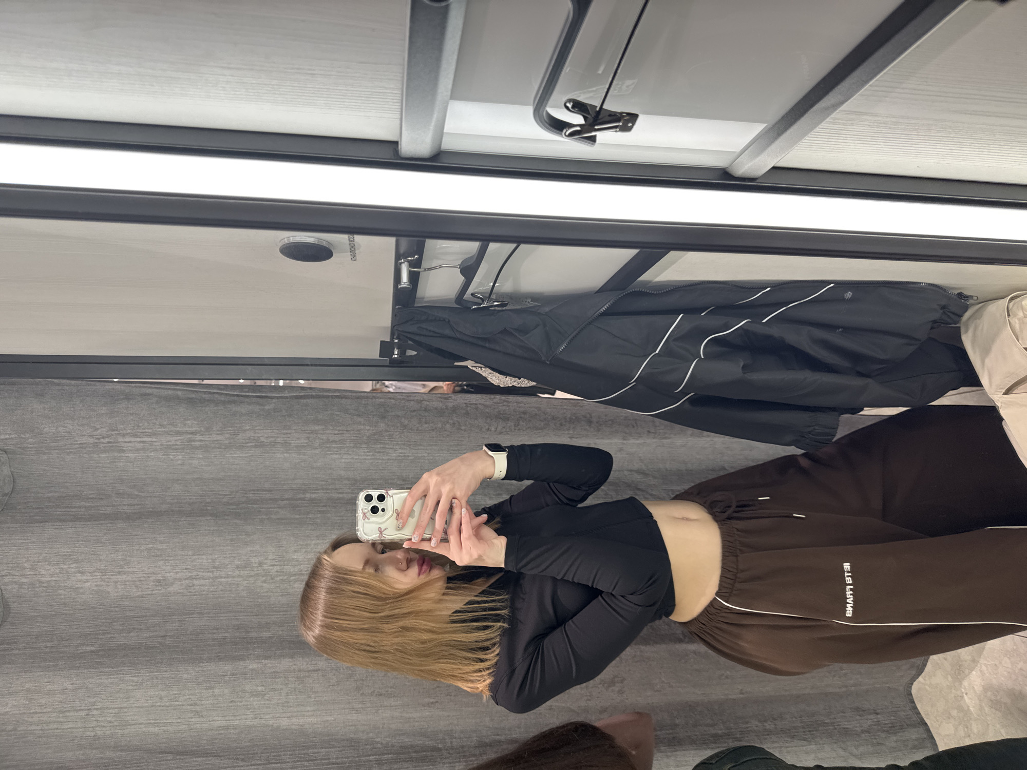 A young woman with long blonde hair is taking a selfie in what appears to be a dressing room. She is wearing a black long-sleeved crop top and brown sweatpants with white trim. She holds a smartphone with a white floral case in her hands. The dressing room has a grey wall behind her and a white ceiling with a black frame and fluorescent lighting. A rack with clothes is visible behind her head. The location in the city of Rostov-on-Don, Russia is not discernible from the image.
