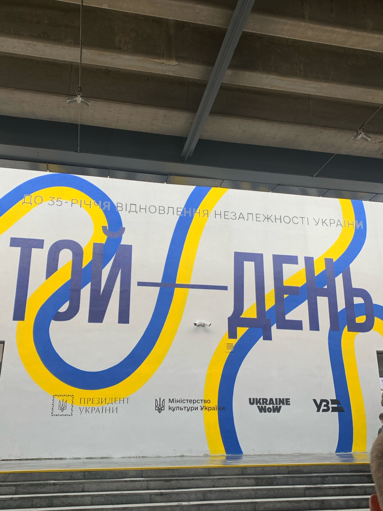 The image shows a mural on the exterior wall of a building in Kyiv, Ukraine. The mural features large, bold lettering in Ukrainian that reads "ТОЙ - ДЕНЬ" (This - Day). The lettering is predominantly blue and yellow, with a blue wave-like design behind it, symbolizing the Ukrainian flag. Above the large text, smaller Ukrainian text states "ДО 35-РІЧЧЯ ВІДНОВЛЕННЯ НЕЗАЛЕЖНОСТІ УКРАЇНИ" (To the 35th anniversary of the restoration of Ukraine's independence). Below the main text, there are several logos, including the coat of arms of Ukraine, the text "ПРЕЗИДЕНТ УКРАЇНИ" (President of Ukraine), "Міністерство культури України" (Ministry of Culture of Ukraine), "UKRAINE WOW," and "УКРІНФОРМ" (Ukrinform), a Ukrainian national news agency. A security camera is visible on the wall to the left of the central text. The mural is positioned above a set of concrete stairs. The surrounding environment appears to be an urban setting, possibly a public space or a government building, given the presence of the presidential and ministerial logos. The overhead structure with visible beams and lighting fixtures suggests an indoor or covered outdoor area, such as a walkway or a transportation hub. No people are clearly visible in the frame, except for a blurred human form at the bottom right edge of the image.
