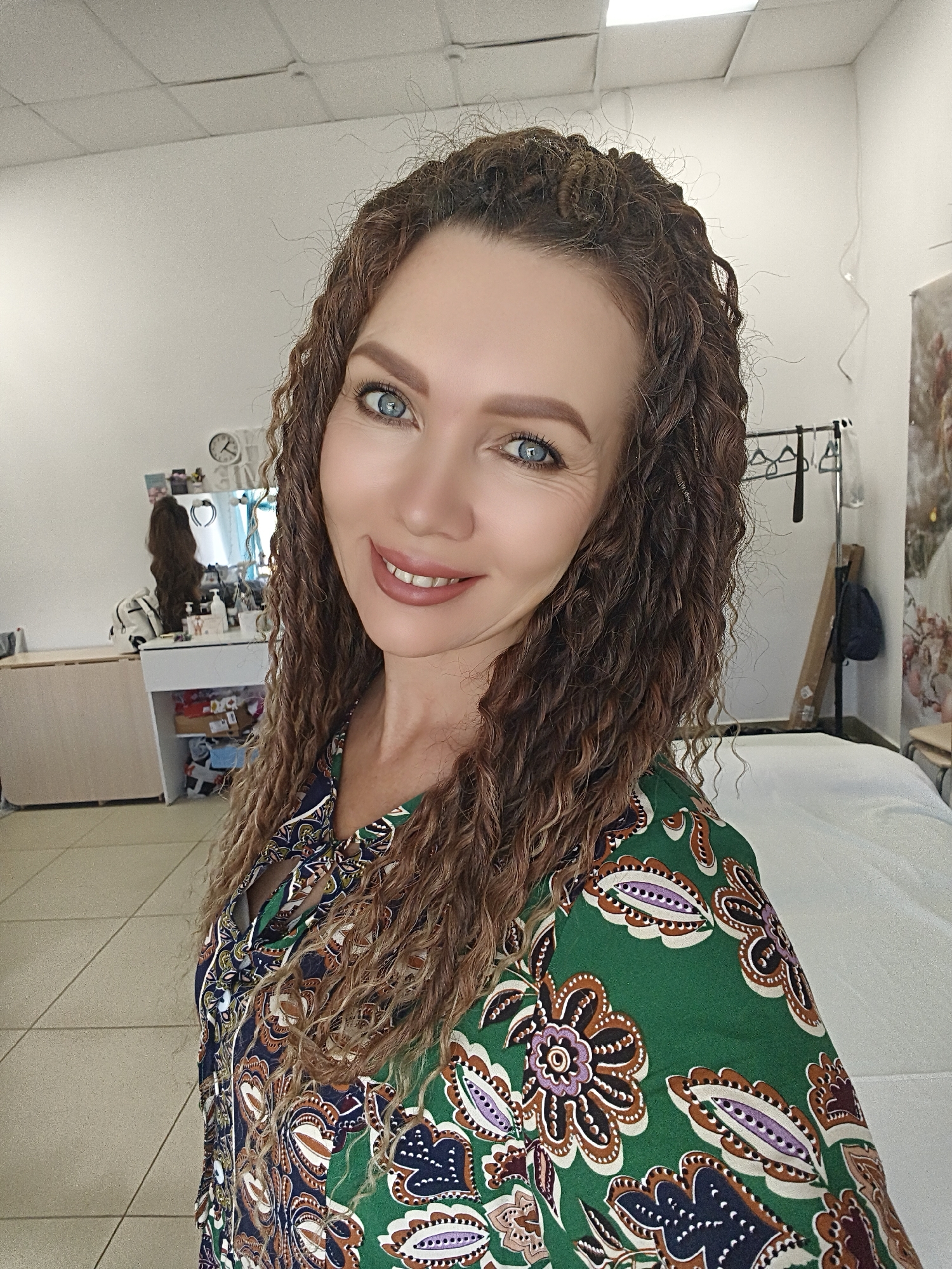 A medium indoor shot of a woman with curly brown hair, blue eyes, and fair skin wearing a green paisley blouse. The background includes a white wall, a dressing table with beauty products, clothing on hangers, and a massage table covered in a white sheet. The city and location are unknown.