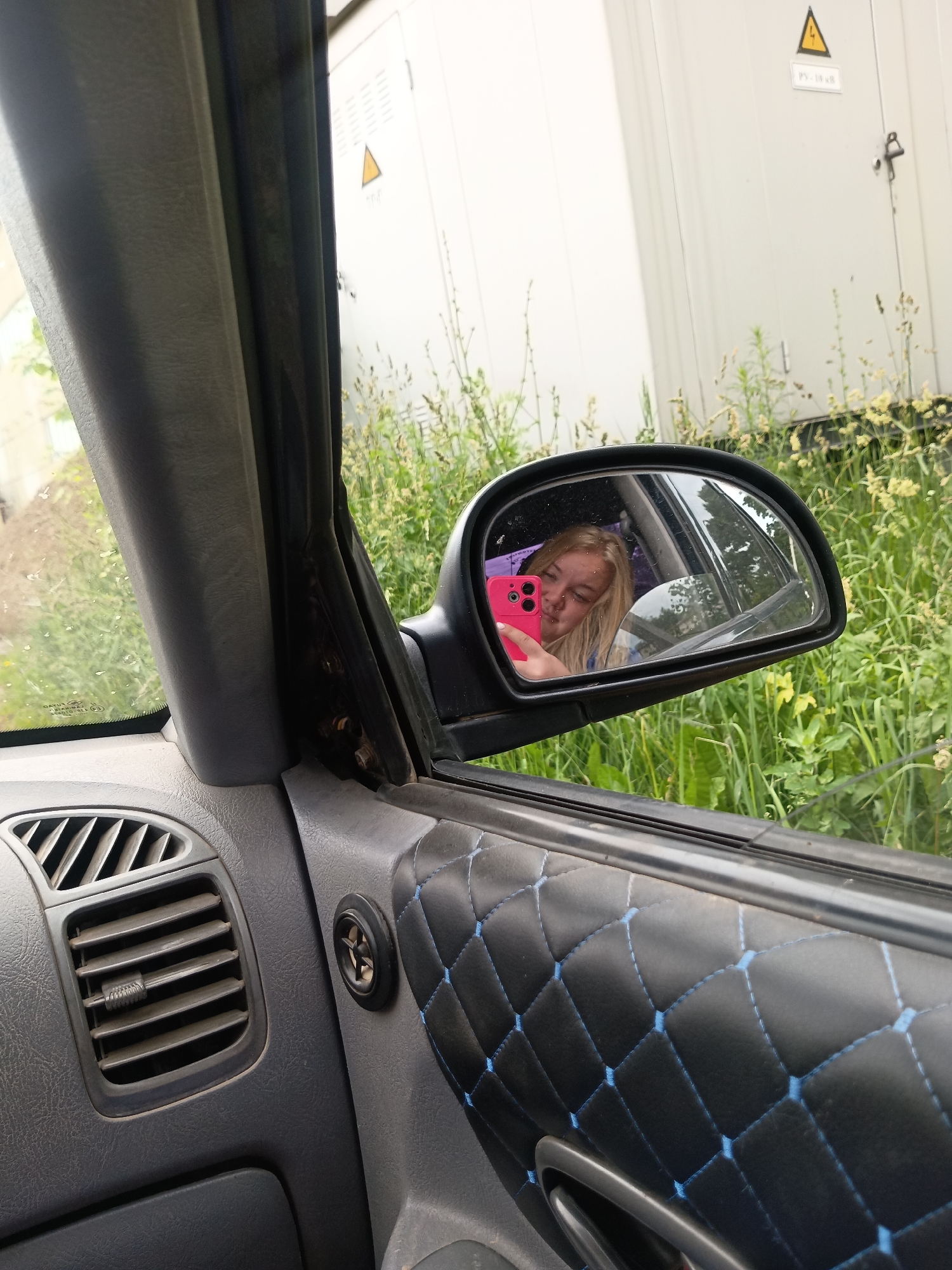 A woman with blonde hair is visible in the reflection of a car's side mirror, taking a selfie with a pink smartphone. She is wearing a blue garment. The car's interior is partially visible, including a gray dashboard with an air vent and a black door panel with diamond-stitched upholstery accented with blue thread. Outside the car, green grass and foliage are present, with a white building in the background. The scene appears to be in the city of Chaikovsky, Russia.