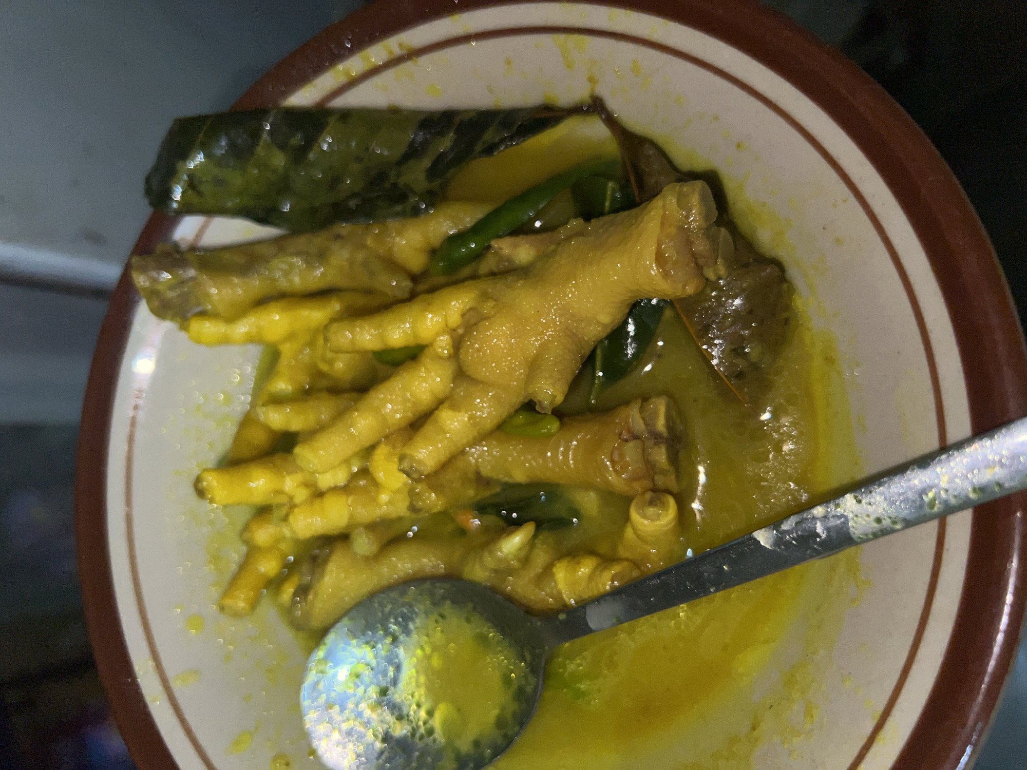 A close-up, overhead shot captures a bowl of cooked chicken feet bathed in a bright yellow broth. A silver spoon rests in the broth, its surface reflecting the surrounding light. The chicken feet, a pale yellow, are arranged somewhat haphazardly in the bowl, with visible toes and joints. Scattered amongst the chicken feet are dark green leaves, likely herbs or spices, and a few small, green chilies. The bowl itself is wide and shallow with a white interior and a distinct brown rim. The background is dark and out of focus, offering no specific environmental context. This image was taken in Klapanunggal, Indonesia.