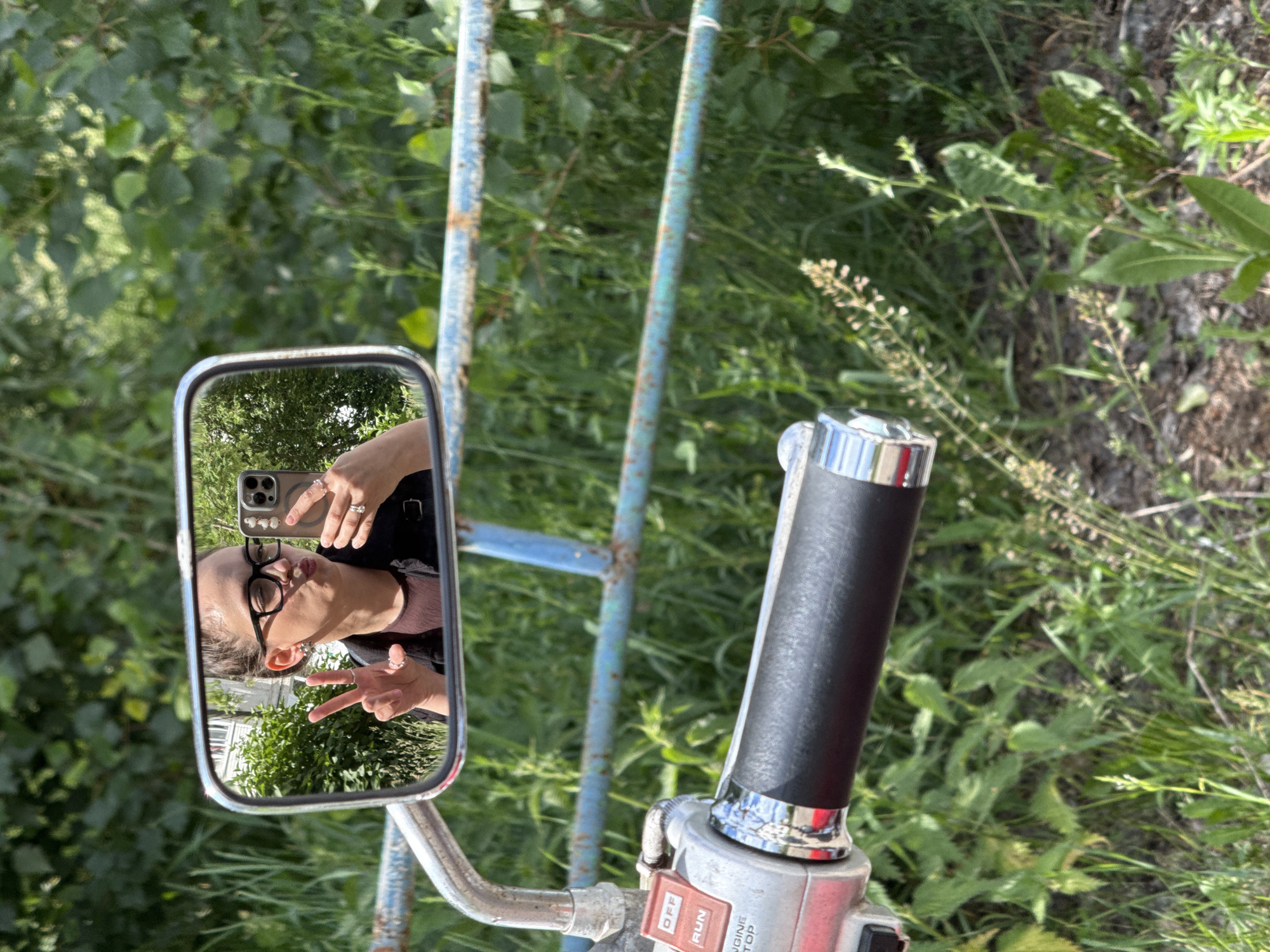 A young woman wearing glasses and a black jacket is taking a selfie in a motorcycle mirror. She is making a peace sign with her left hand and holding a smartphone in her right hand. The mirror reflects a building and greenery. The motorcycle's handlebar, a red and silver switch, and a blue metal fence are visible in the foreground. The background consists of lush green foliage.