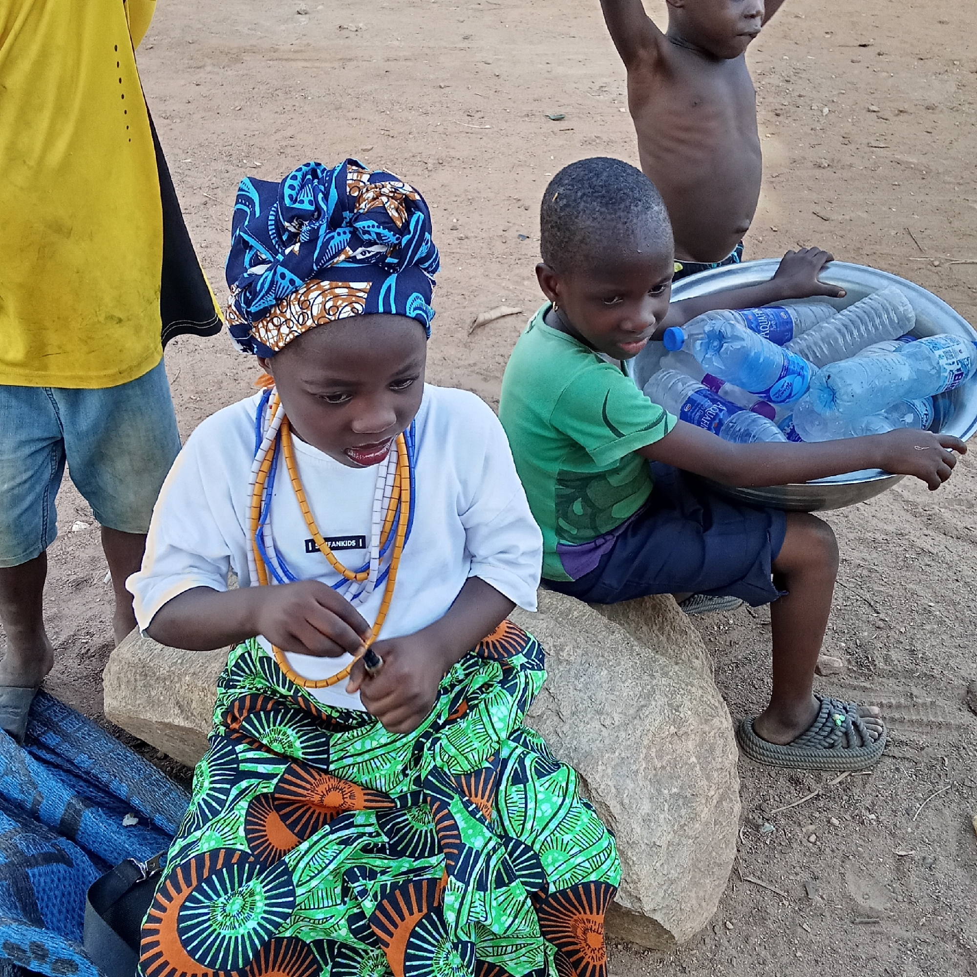 This outdoor image from Akwanga, Nigeria, captures a group of four children on a dusty, earthen ground, likely in the late afternoon or early evening given the soft, warm light.
In the foreground, a young girl is seated on a large, smooth rock. She wears a vibrant blue, white, and brown patterned headwrap, a white long-sleeved t-shirt, and a colorful green, orange, and black patterned skirt. Several multi-colored beaded necklaces (orange, white, and blue) are around her neck, and she appears to be holding a small object or manipulating beads with her hands. Visible text on her t-shirt reads "I FANKIDS".
To her right, a boy is seated on the ground, wearing a green t-shirt and dark shorts, with gray sandals on his feet. He holds a large, silver metal basin filled with numerous plastic water bottles, some clearly bearing the "Aquafina" brand label. His gaze is directed towards the right.
Behind him, another young boy stands shirtless, with his arms raised above his head. On the far left, partially visible, is another child wearing a yellow t-shirt and light blue denim shorts, standing barefoot.
The ground is uneven and dry. A blue tarp or fabric lies on the ground to the left. The children's expressions are generally neutral or focused on their activities, suggesting a quiet, everyday moment. The presence of water bottles implies possible trade or distribution.