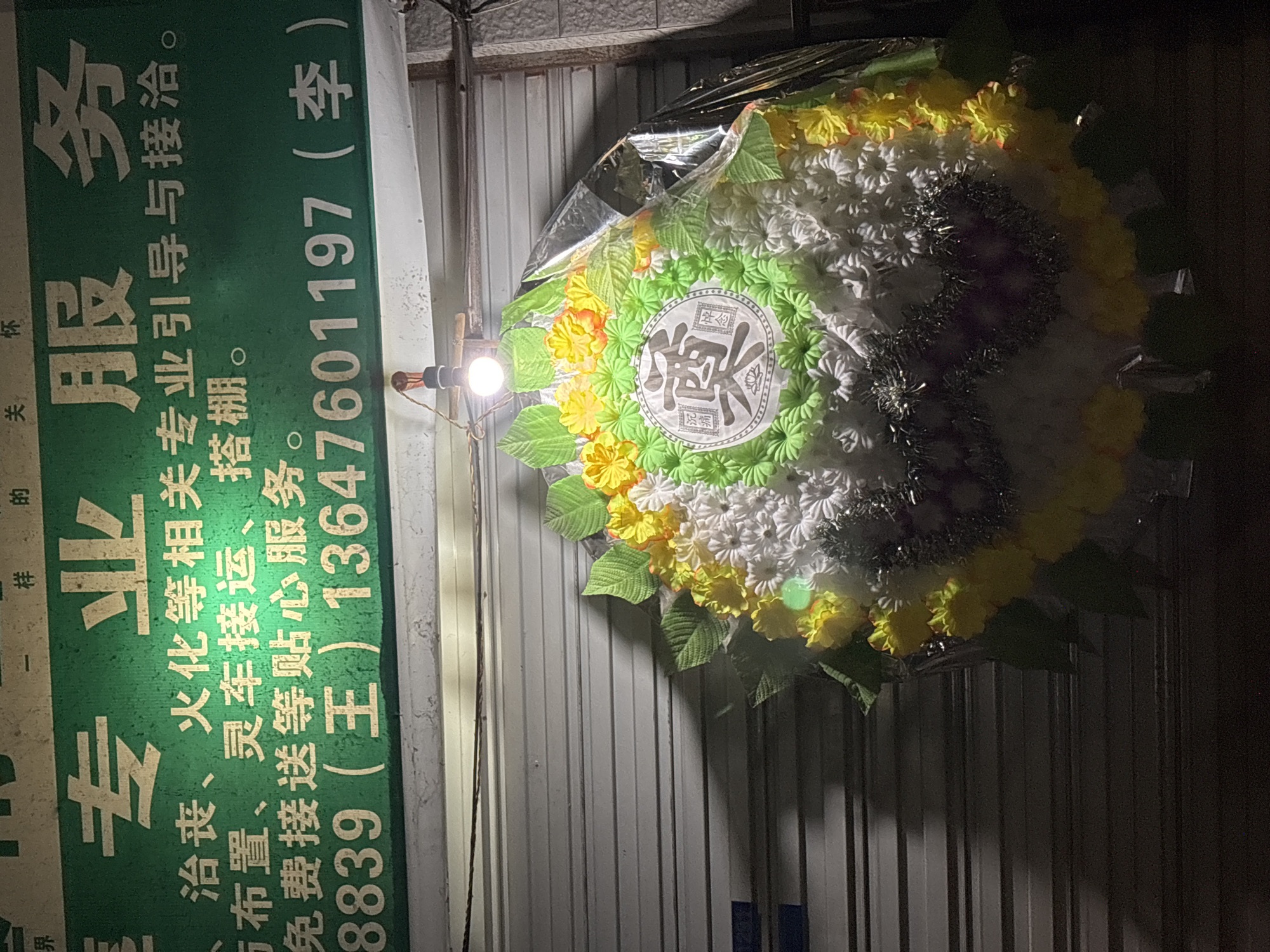 Here is a description of the image:
The image shows a business sign next to a floral wreath, possibly related to funeral services. The sign is green with white Chinese characters advertising "Professional Services", listing services like funeral arrangements, cremation, transportation, and free pick-up. A phone number is also visible on the sign. The floral wreath is made of white, yellow, and green artificial flowers with a central emblem. The background features a corrugated metal surface. The setting is likely an urban area in China, based on the language and type of business. It is not possible to confirm the city or district from the image.
