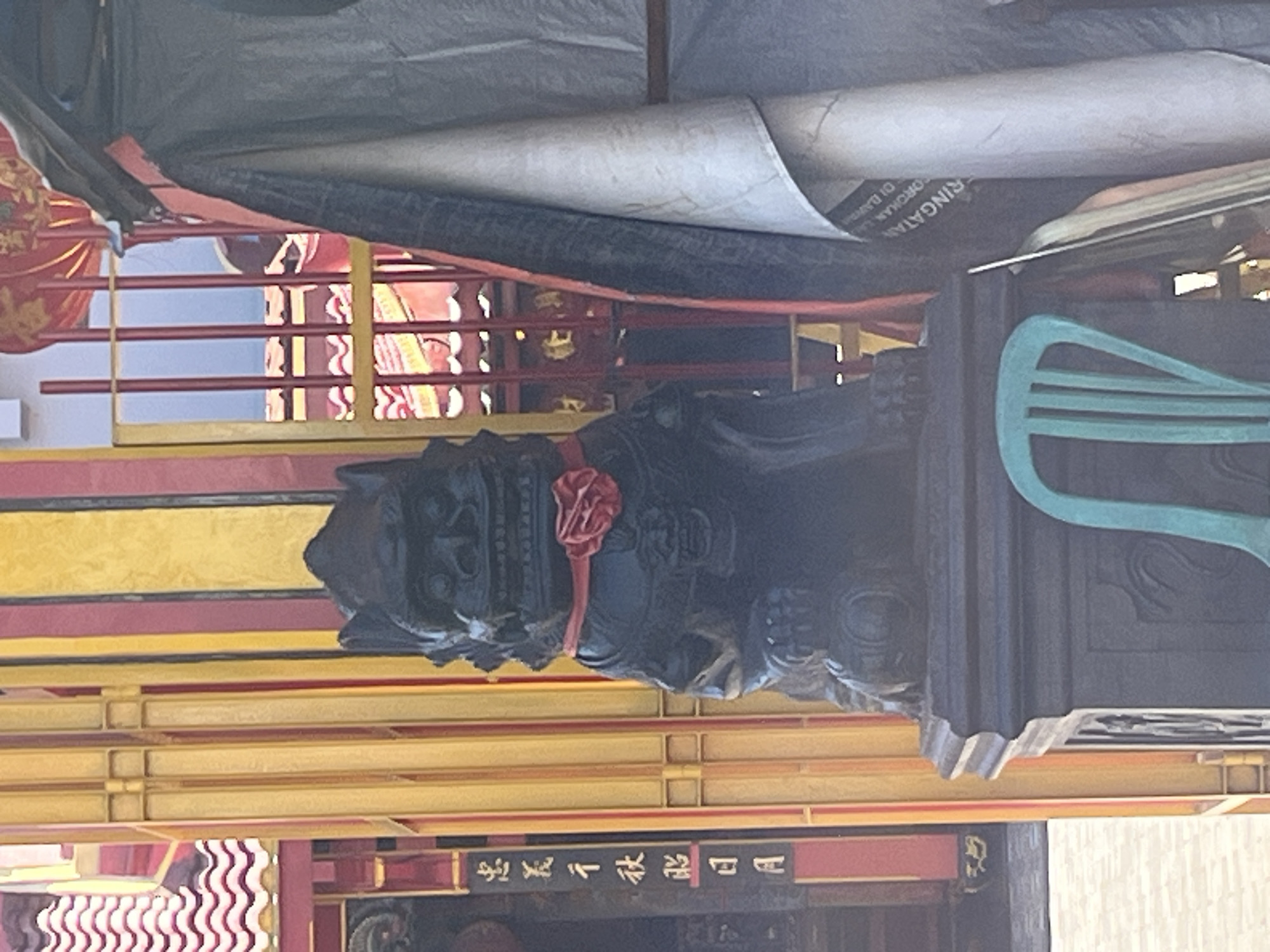 In Desa Tambakreja, Indonesia, a black stone statue of a lion-like creature, possibly a guardian or temple lion, stands prominently on a dark pedestal. The statue is adorned with a red collar that has a red floral embellishment. Behind the statue, a vibrant yellow and red structure with vertical bars is visible, suggesting it might be part of a temple or traditional building. A light-colored rolled-up tarp or fabric is draped to the right of the statue, obscuring a portion of the background. To the left of the statue, a black column features vertical script in white, possibly Chinese or other Asian characters. A bright turquoise chair is placed in front of the pedestal, partially obscuring it from view. The ground in front of the structure appears to be paved with light-colored square tiles. The overall scene suggests a cultural or religious site within the village.