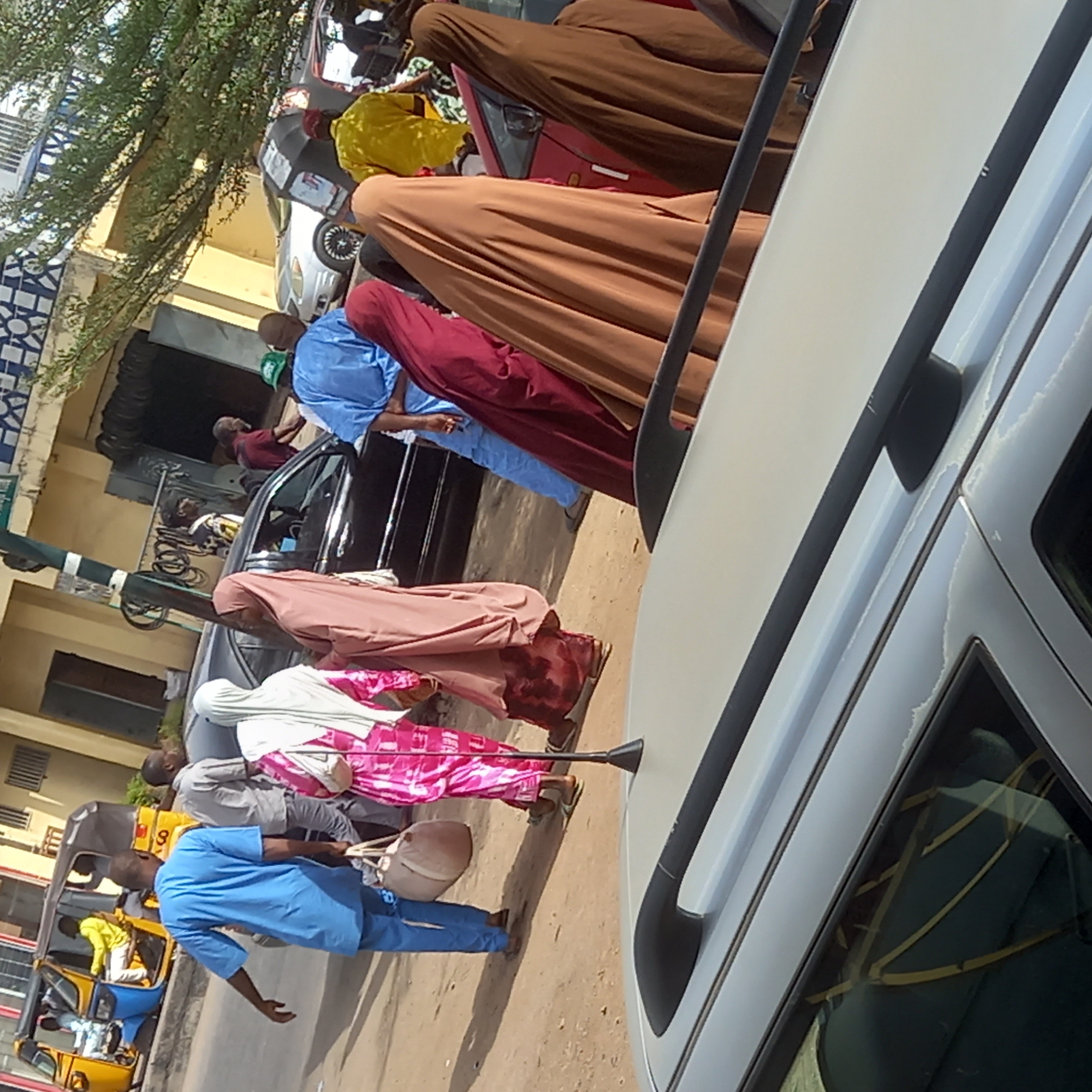This image captures a vibrant street scene in Sardauna, Nigeria, under bright daylight. The perspective is taken from next to a light-colored vehicle, whose side and black roof rails dominate the right foreground.
Numerous people, predominantly dressed in traditional attire, are visible. Several women are seen wearing long, flowing garments and head coverings: one in brown, another in maroon, and a third in a striking pink and white patterned dress paired with a white headscarf, who is using a black walking stick. A man in a light blue tunic and trousers walks towards the left, carrying a beige bag over his shoulder. Another man in a grey shirt and pants stands behind him.
The setting appears to be a busy street or marketplace. A yellow and blue Keke Napep (auto-rickshaw) with the number "9" prominently displayed on its side is visible on the far left, with passengers inside. Other cars, including a dark-colored one, are present along the dusty road. In the background, buildings with yellow facades and some featuring blue and white patterned designs line the street. Green foliage from trees overhangs the left side of the scene, partially obscuring the buildings. A green and white painted pole is also visible. The clear sunlight and visible shadows suggest it is daytime. The image depicts daily life and movement within an urban or semi-urban Nigerian environment.