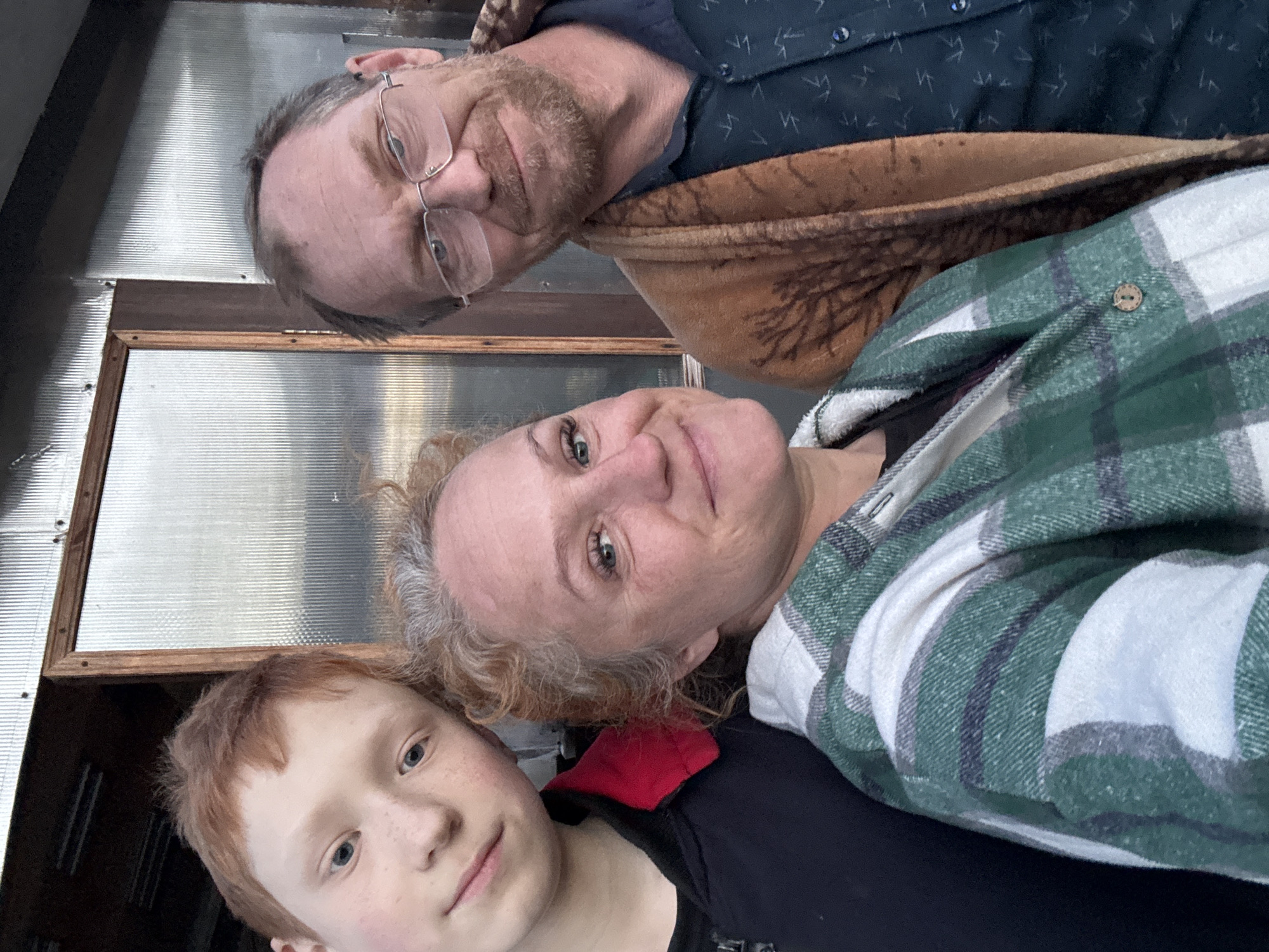 A selfie taken outdoors depicts three people: a red-haired child in a black t-shirt and red garment, a middle-aged woman with curly, graying hair wearing a green and white plaid shirt, and a middle-aged man with glasses wearing a blue patterned shirt and brown jacket. They are positioned close together. The background shows a corrugated metal wall or structure with a framed window. The location is implied to be in the city of Chaikovsky, Russia, based on the user's prompt.