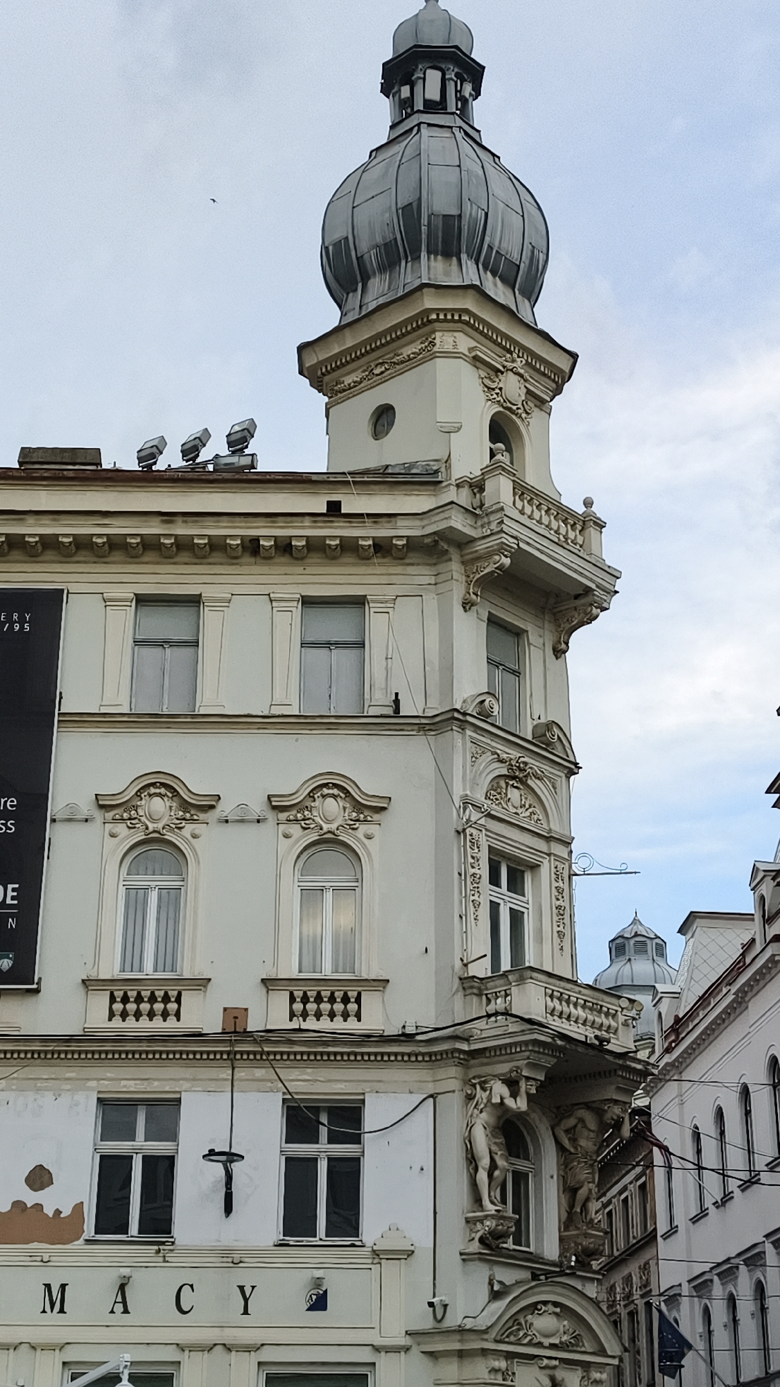 The image shows a detailed view of the exterior of a building in Sarajevo, Bosnia and Herzegovina. The building features ornate architectural elements, including a prominent domed tower at the top, decorative moldings, arched windows, and statues integrated into the facade. The building's facade is predominantly white with cream-colored accents. On the lower part of the building, the word "MACY" is visible, suggesting it might be a commercial establishment. There are windows on multiple levels, some with intricate decorative patterns above them. A black banner with white text is partially visible on the left side of the building. The sky is overcast with a few faint clouds. The overall impression is of an old, architecturally rich building in an urban setting.