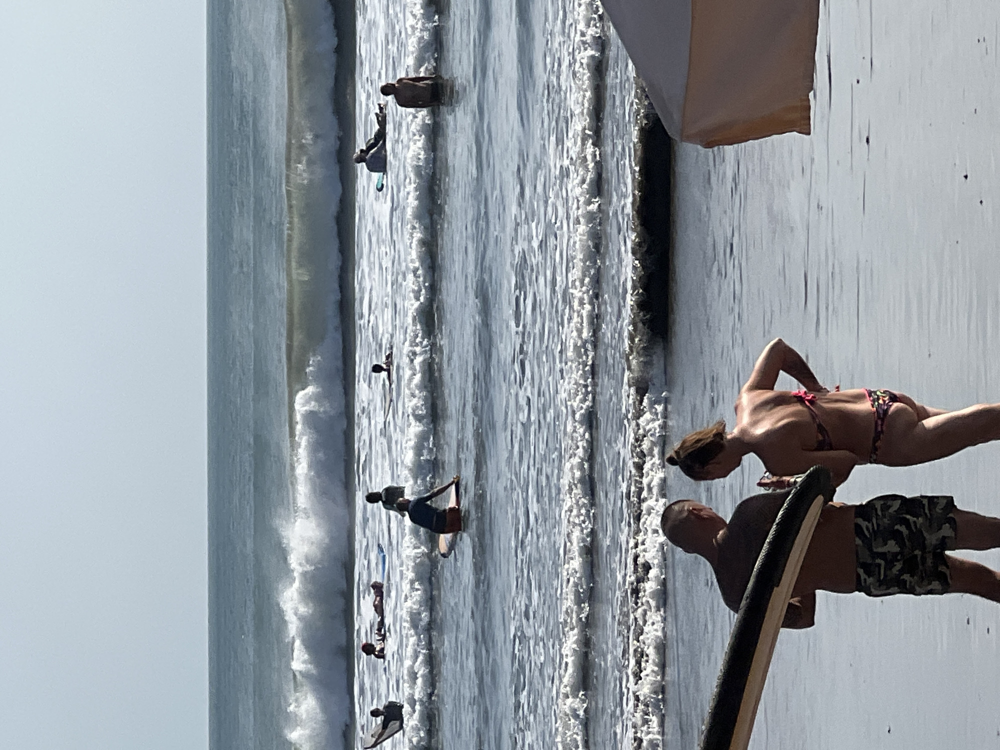 In Kuta, Indonesia, on a sunny day, several people are enjoying the ocean. In the foreground, a man and a woman stand on the wet sand, facing each other and appearing to be in conversation. The man is holding a surfboard. The woman is wearing a bikini. Behind them, the ocean has moderate waves breaking. Several surfers are visible in the water, some paddling on surfboards, others sitting on them, and one person standing in the water near the shore. The horizon line is clear, with a light blue sky above the ocean. The overall scene depicts a typical day at the beach with recreational activities.