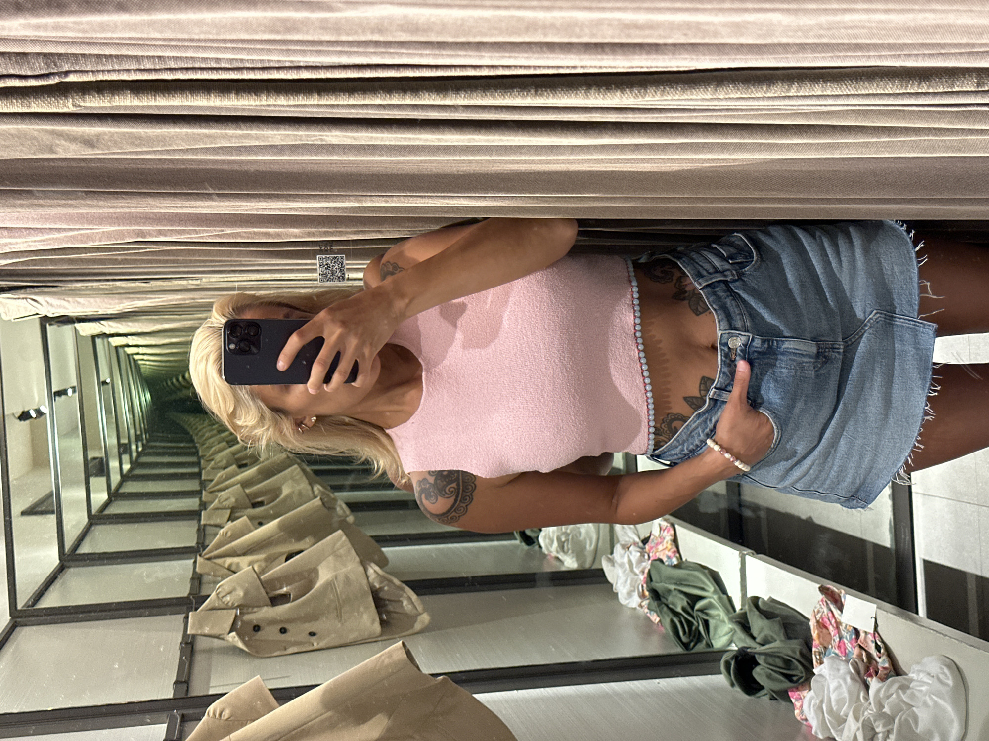 The image shows a woman taking a selfie in a fitting room. She has blonde hair and is wearing a pink crop top and a denim skirt. Her left arm is bent at the elbow, with her hand resting on her hip. She has tattoos on her left arm and hip. The fitting room has mirrored walls that create a sense of infinite depth, reflecting rows of mannequins or displays of clothing. The lighting is artificial, suggesting an indoor setting. Some clothing items, including a tan jacket and several crumpled tops, are visible in the reflection. The overall impression is one of a casual shopping experience. There is a QR code visible on the wall in the background. The image appears to be taken during the day, given the brightness of the artificial lights.