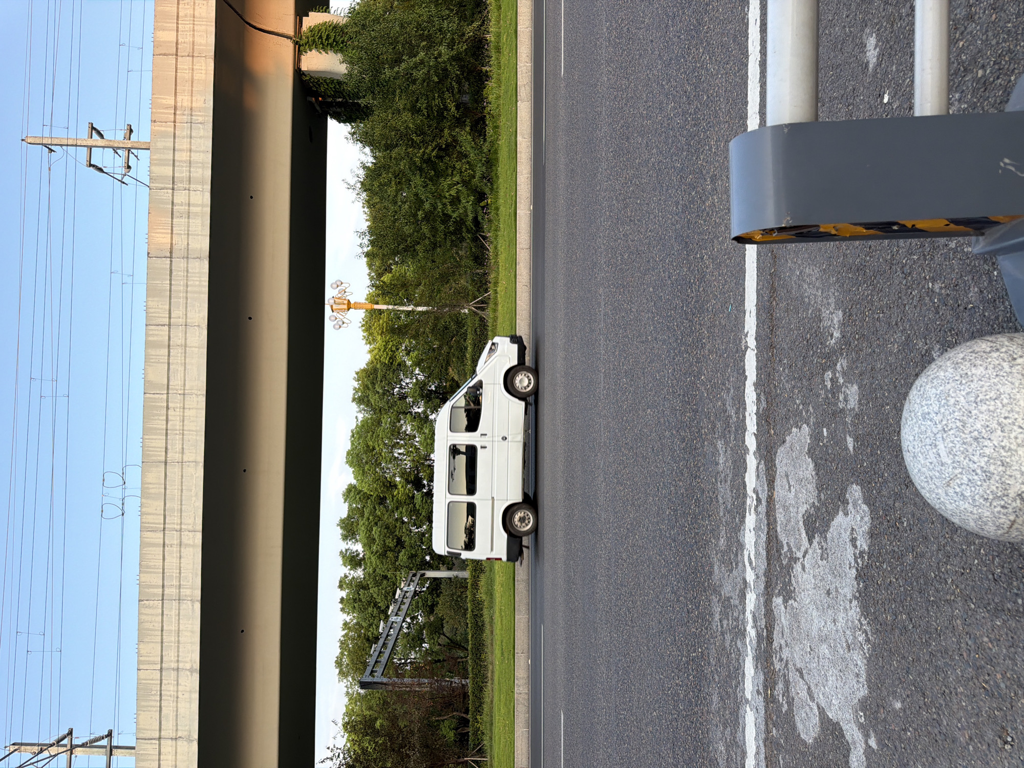 A white van is traveling on a road in Jiangjin, China. The van is white, with tinted windows and four visible wheels. It is positioned in the right lane of a multi-lane road. Above the road, a concrete overpass spans across the image, supported by pillars. Power lines are visible above the overpass, suspended from poles. To the left of the road, there is a grassy area with trees and a lamppost. The sky is clear and blue, indicating daytime. The foreground of the image shows the asphalt road with white lane markings and some scattered white material. A grey concrete bollard and a section of a guardrail are also present in the foreground.