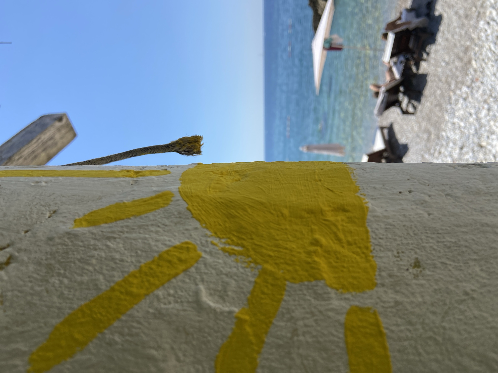 This is a vertical, eye-level, medium shot photograph of a painted handprint on a textured white pillar in the foreground. The handprint is painted in a bright yellow and appears to be in the process of drying, with visible brushstrokes. To the right of the pillar, a frayed rope hangs downwards. Beyond the pillar, a bright blue sky dominates the upper right portion of the image. Below the sky is the clear blue sea, with a visible horizon. The middle ground shows a pebbly beach with several lounge chairs and umbrellas. Two people are visible, partially obscured by lounge chairs, relaxing on the beach. The beach is a part of Prijevor, Montenegro.