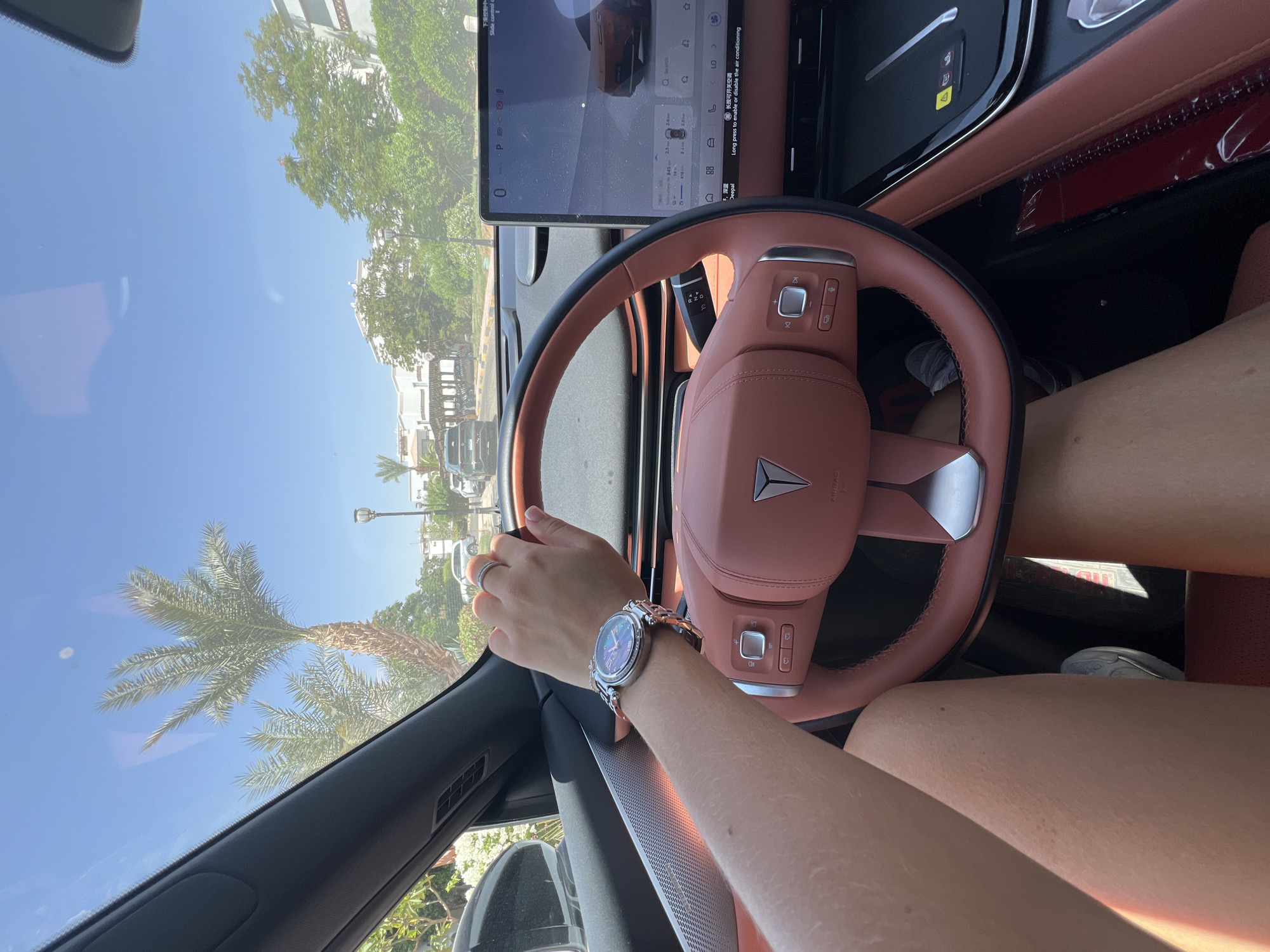 In an interior shot from a vehicle's perspective, a person's hands and legs are visible, suggesting they are driving. The driver's left hand, adorned with a watch and a ring, rests on the steering wheel. The steering wheel itself is a reddish-brown leather, centrally featuring a triangular logo. The dashboard includes a large touchscreen display showing vehicle information, with text in Chinese characters and English prompts. The car's interior appears to be luxurious, with similar reddish-brown leather accents. Through the windshield, a sunny, outdoor scene is visible, featuring palm trees, white buildings with arched windows, and other vehicles. The environment suggests a warm, possibly tropical or desert climate. The presence of palm trees and the architectural style of the buildings are consistent with locations like Sharm El Sheikh, Egypt, known for its resorts and arid landscape. The bright sunlight and clear blue sky further reinforce this impression.