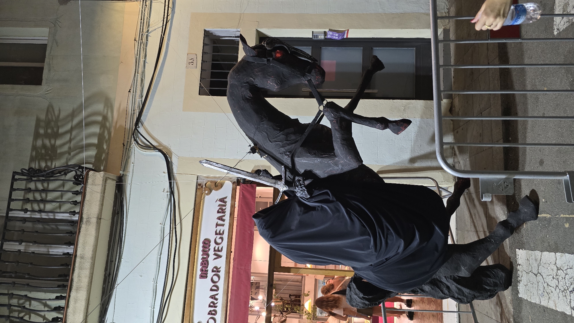 This is an outdoor, eye-level, medium shot photograph taken at night in Barcelona, Spain. The image features a large, dark sculpture of a horse with a rider cloaked in black, as if to represent the "Ringwraith" from The Lord of the Rings. The horse is rearing up on its hind legs, and the rider is holding a sword. The sculpture is positioned in front of a building with a sign reading "NABUCCO OBRADOR VEGETARIA" and a red awning, suggesting it is a shop. A balcony with ornate metal railings is visible above the shop. The ground is paved, and a metal barricade is in the foreground, suggesting a street or public space. In the background, a door with a number "33" above it is visible, and two women are partially visible through the shop window. The overall lighting suggests artificial illumination from streetlights and shop interiors.