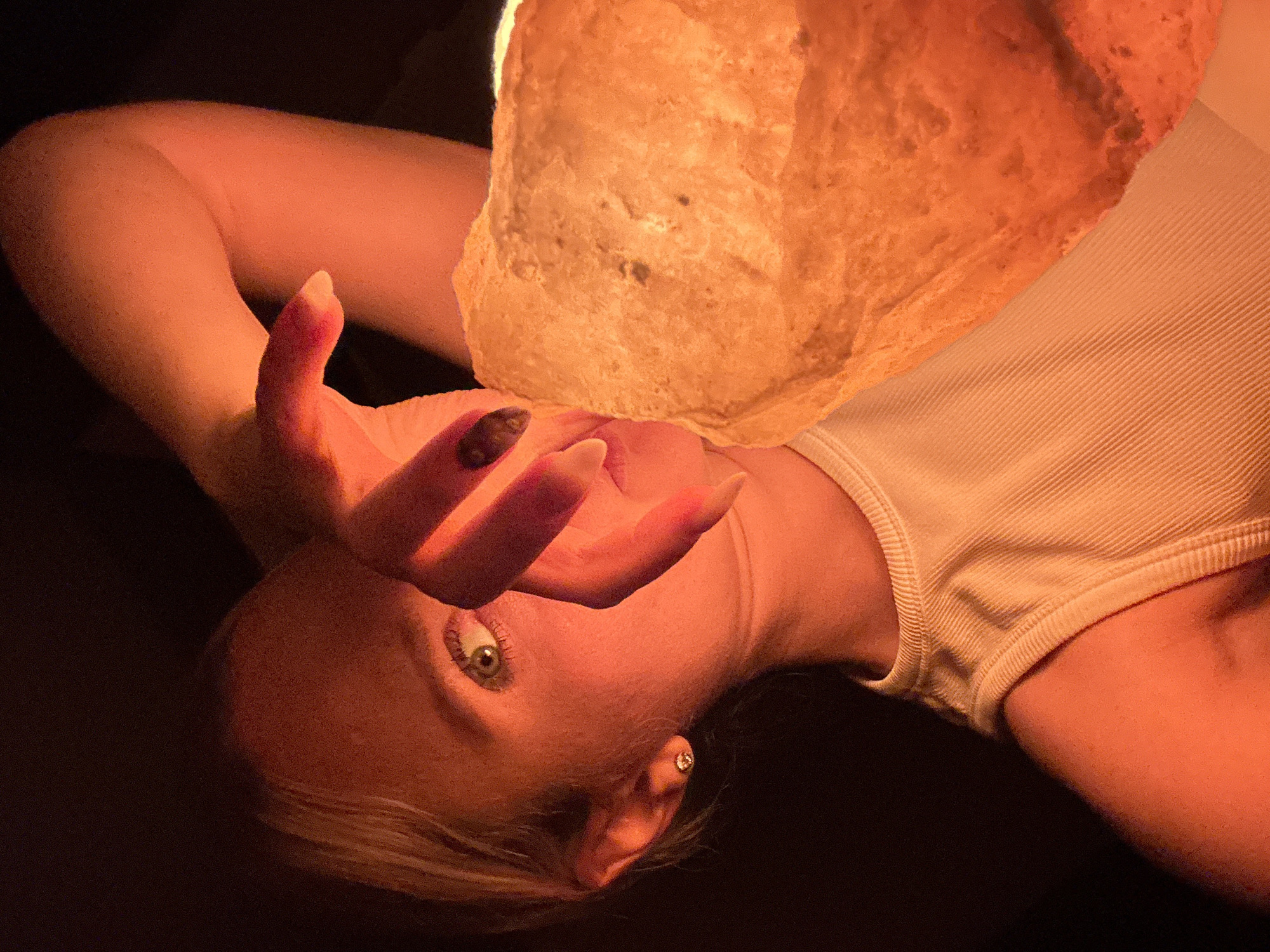 A person lies on a dark surface, angled towards the viewer. The person has light skin and appears to be female, with blonde hair. She wears a light-colored, ribbed tank top. A hand with dark polished nails is held up near her face. A large, illuminated object, possibly a salt lamp, is positioned above her face. The location is not discernible and cannot be determined to be in Esenyurt, Turkey, from the image alone.
