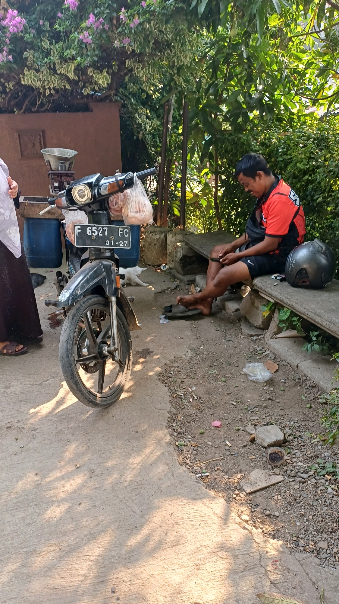 A middle-aged man in an orange shirt and black shorts sits on a concrete bench, looking down at his phone. A black motorcycle is parked to his left, with two plastic bags of goods hanging from the handlebars. A grey motorcycle helmet rests on the bench next to him, and a white cat is lying on the ground near the motorcycle. The scene appears to be outdoors in front of a building, with a wall and foliage visible in the background. The ground is a mixture of dirt and gravel, with some scattered litter. The image is taken during the day, with sunlight casting shadows on the ground. The location is identified as Gunung Putri, Indonesia.