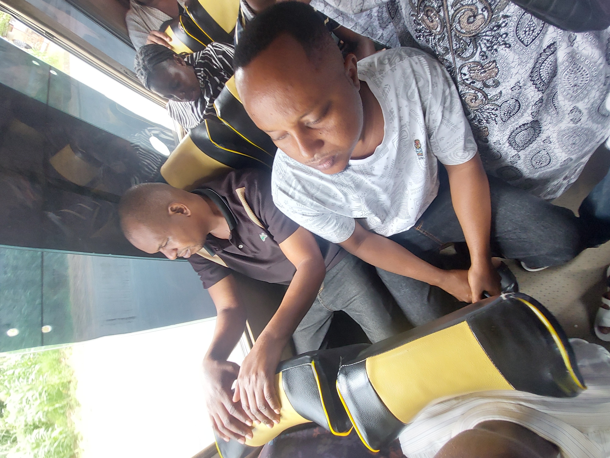 A group of people is seated inside a vehicle, possibly a bus or van, with at least two men visible in the foreground. One man is wearing a dark polo shirt and the other a patterned white t-shirt. Both are wearing jeans. The man in the t-shirt appears to be asleep or resting his eyes. They are seated next to each other, with their hands resting on the back of the seat in front of them, which is black and yellow. Other people are visible in the background, including someone wearing a striped shirt and another in a white patterned garment. Through the vehicle window, some greenery can be seen. It is likely that this is Dar es-Salaam, Tanzania, but that cannot be confirmed with the image alone.
