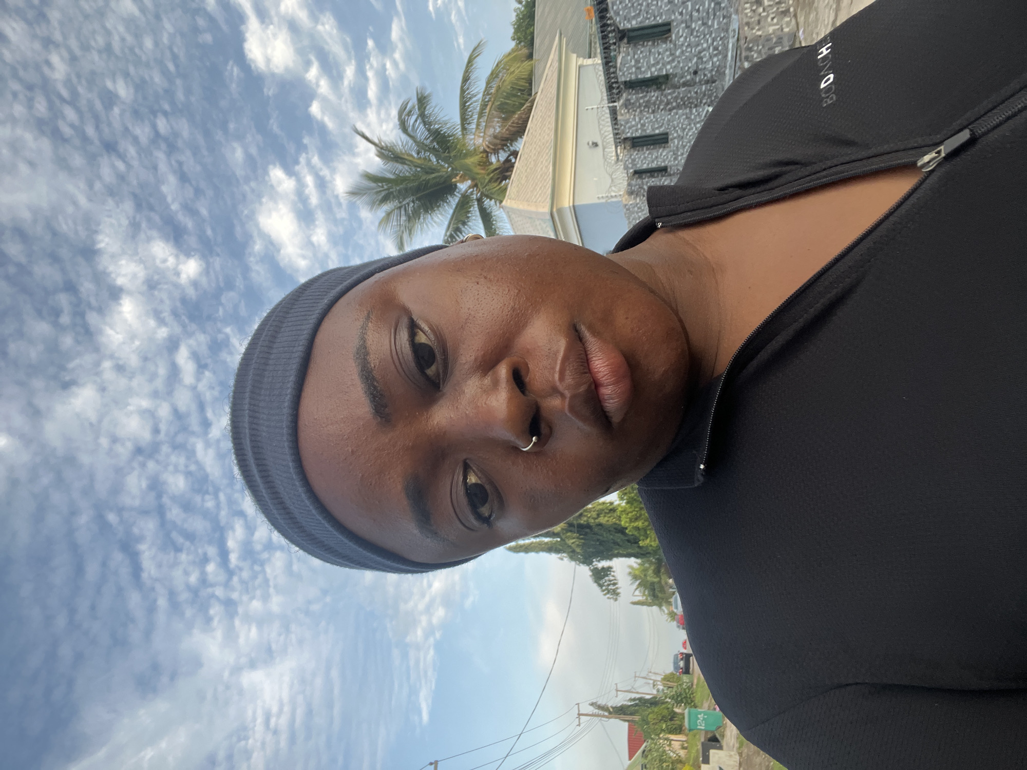 A medium shot shows a dark-skinned woman wearing a black athletic zip-up jacket and a dark gray headband. She has a nose ring. The background is an outdoor scene with a cloudy sky, palm trees, houses, streetlights, and a few cars. The scene appears to be in a residential area in Abuja, Nigeria. The lighting suggests it is either early morning or late afternoon.