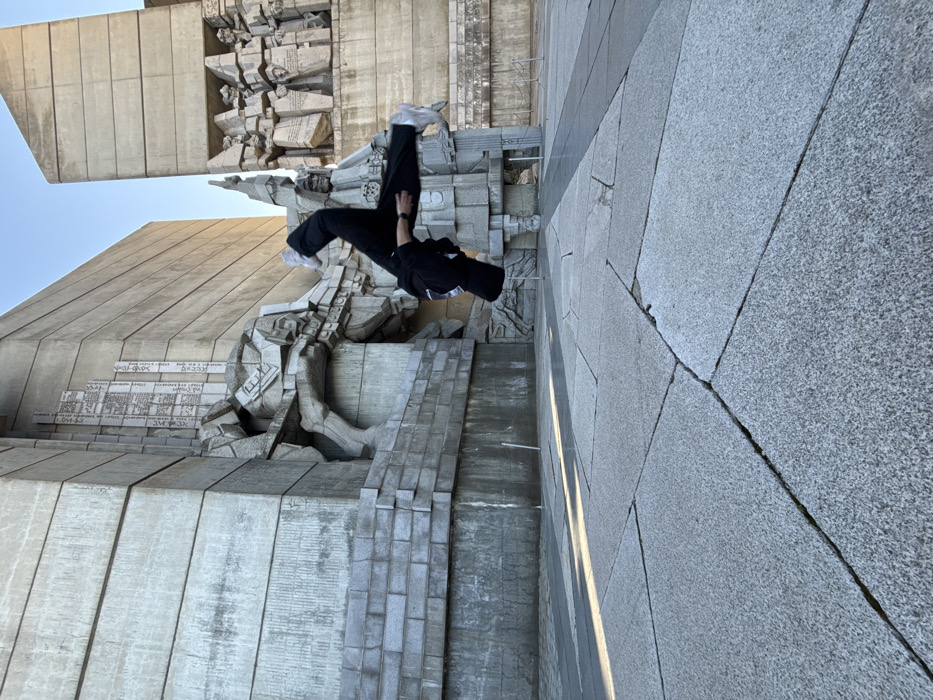 An individual is captured mid-air during an acrobatic flip or jump, positioned horizontally against the backdrop of the Monument to 1300 Years of Bulgaria in Shumen, Bulgaria. The person is dressed in black pants, a black long-sleeved top, and white sneakers, with a wristwatch visible on the left wrist. The monument, constructed from large, grey concrete blocks, features prominent brutalist-style sculptures including a figure seemingly breaking free from a wall on the left, a central figure with an upraised arm, and a relief depicting multiple figures on the right. The ground is paved with large, light grey granite slabs, displaying visible seams and minor cracks. Sunlight casts distinct diagonal shadows across the pavement. The sky above is clear and light blue.